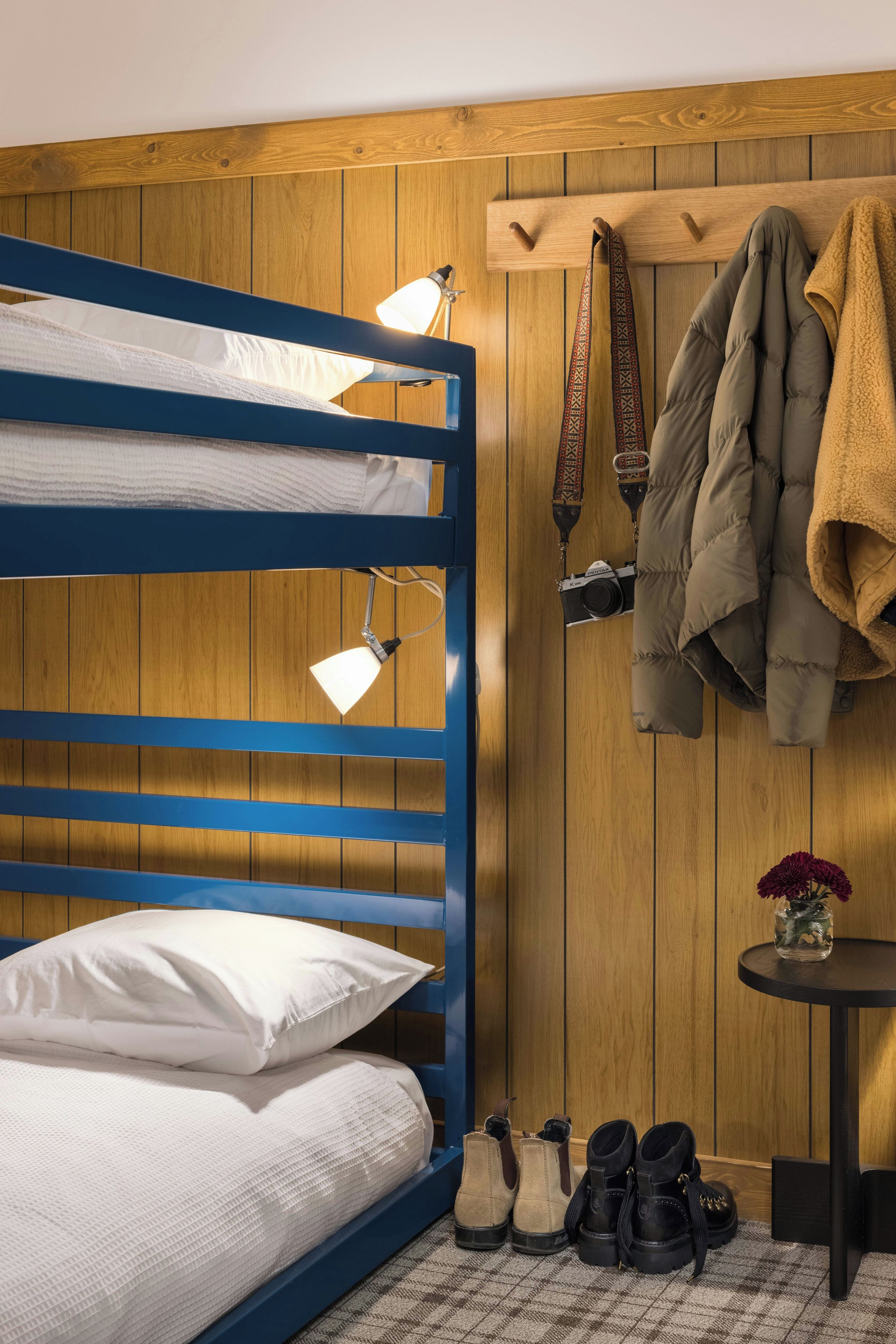 A bunk bed with two lamps, a coat rack with jackets, a camera, and a small table with a vase.