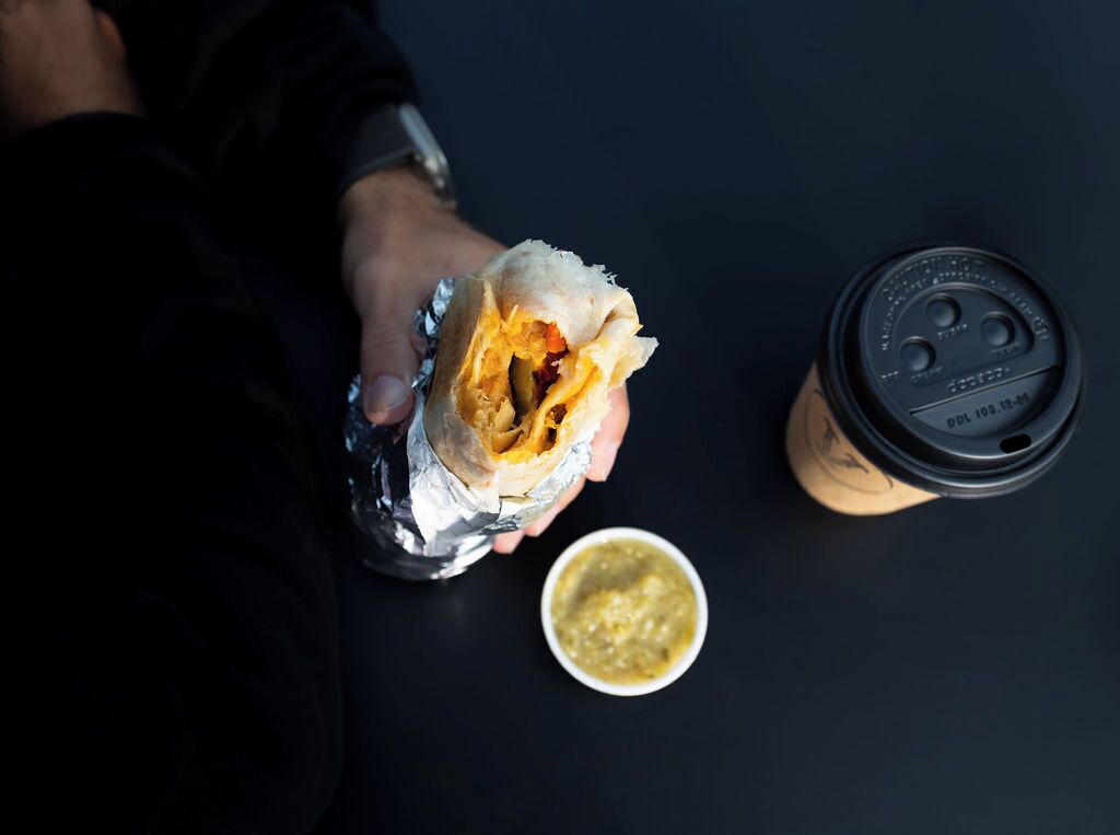 A burrito wrapped in foil, a cup with a lid, and a small bowl of sauce on a dark surface.