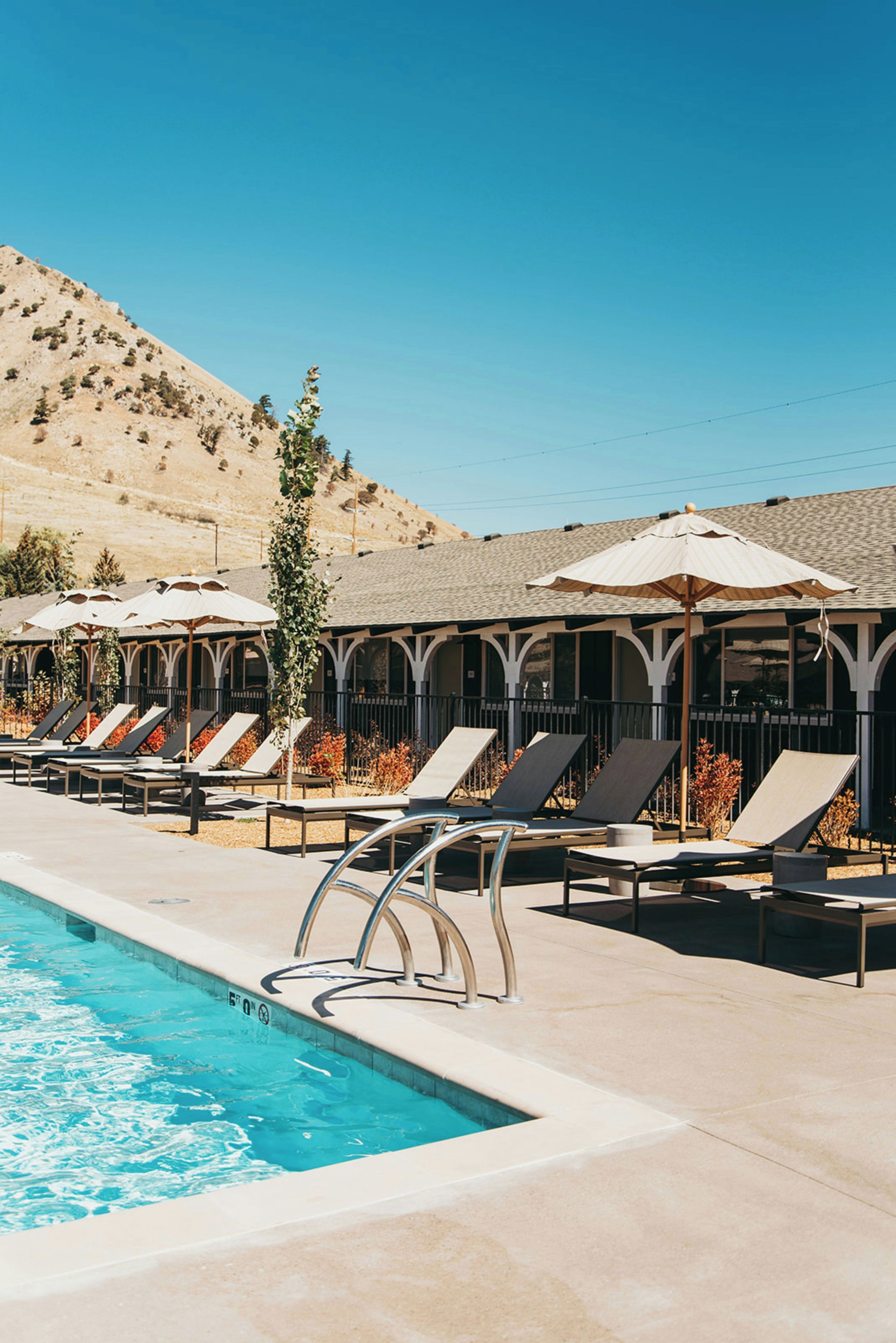 A poolside area with lounge chairs and umbrellas, a clear sky, and a hilly landscape in the background.