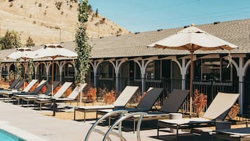 A poolside area with lounge chairs and umbrellas, a clear sky, and a hilly landscape in the background.