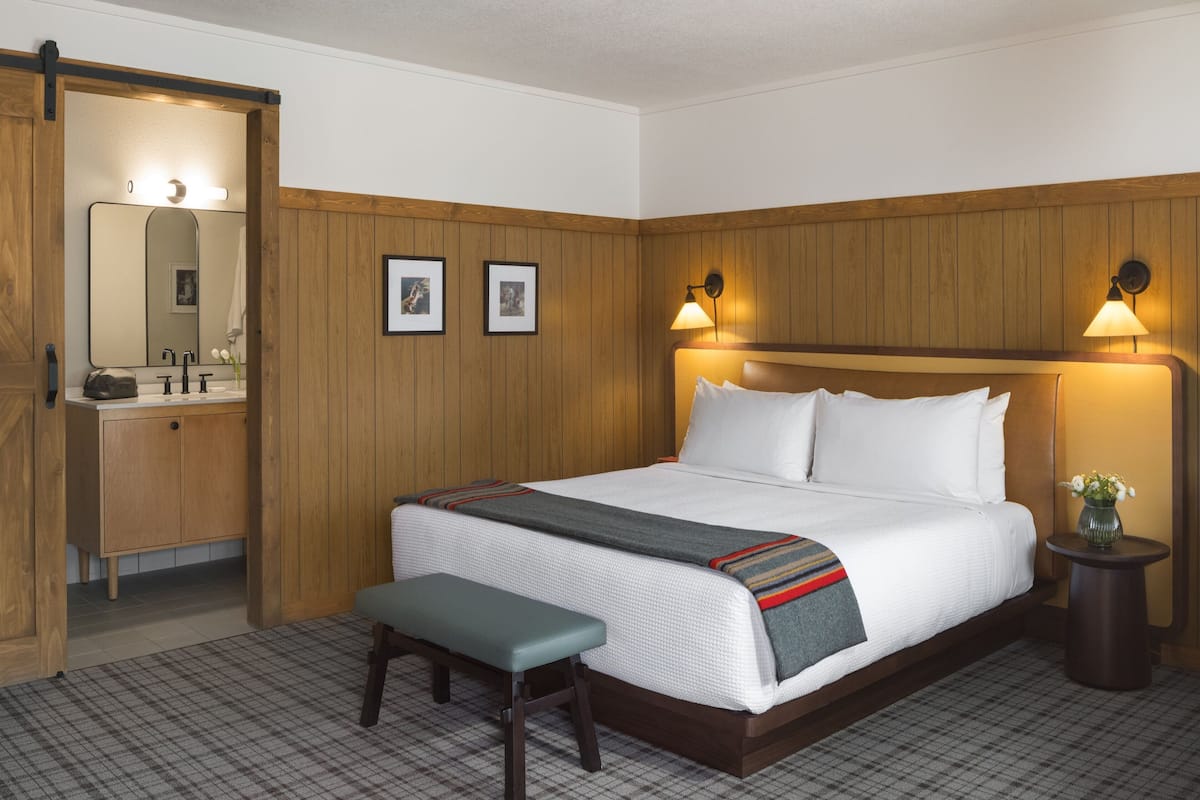 A hotel room with a wooden paneled wall, a bed with white linens, a bench, a small table with a vase, and a bathroom with a mirror and sink.