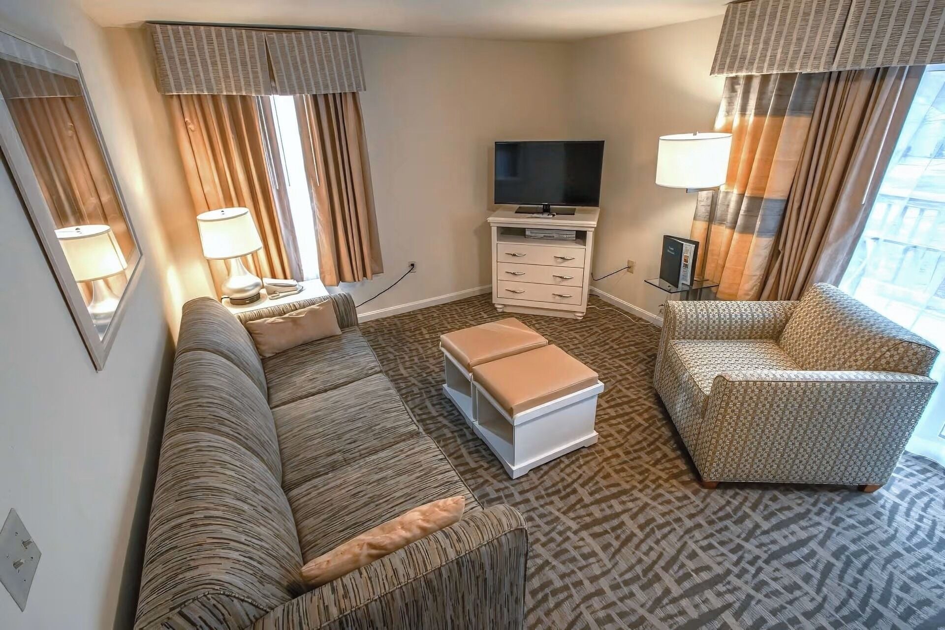 Ett hotellrum med en soffa, fåtölj, pall, TV och ett litet bord med lampor.