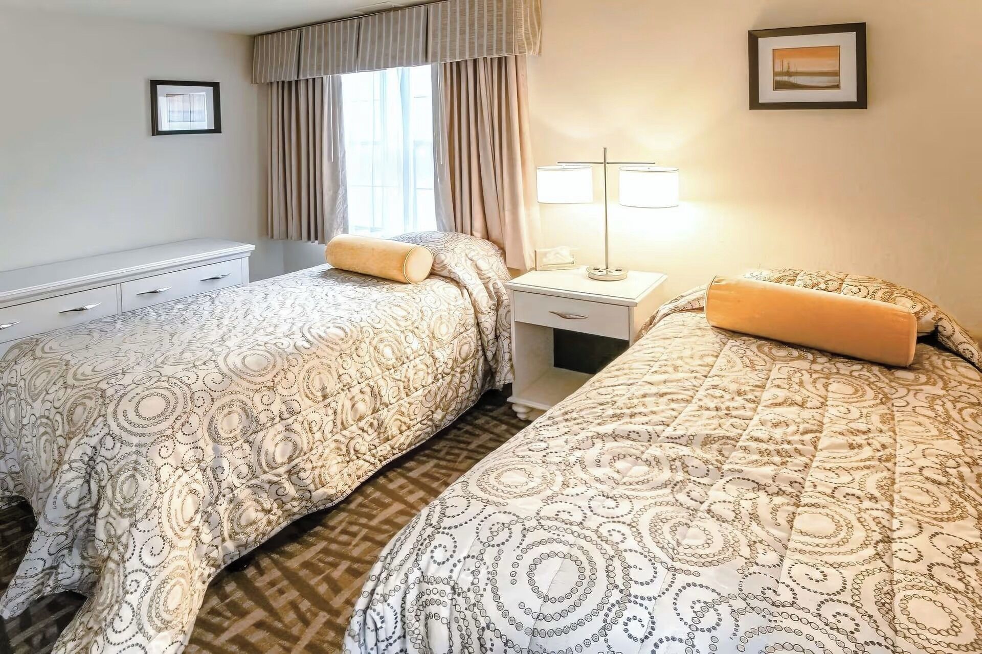 Een hotelkamer met twee bedden, een nachtkastje met een lamp, een raam met gordijnen en een ingelijst schilderij aan de muur.