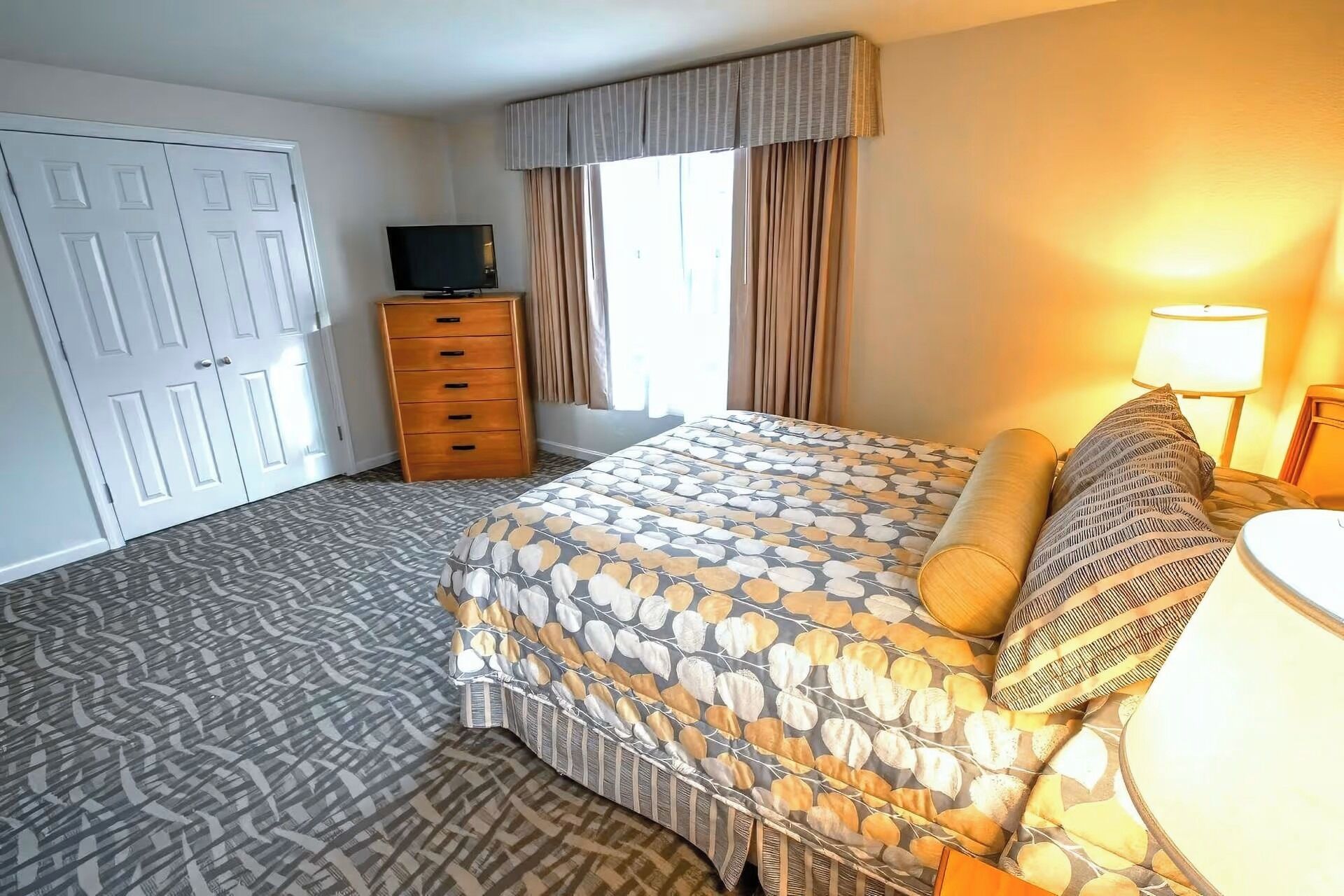 Habitación de hotel con cama, cómoda, televisor y ventana con cortinas.