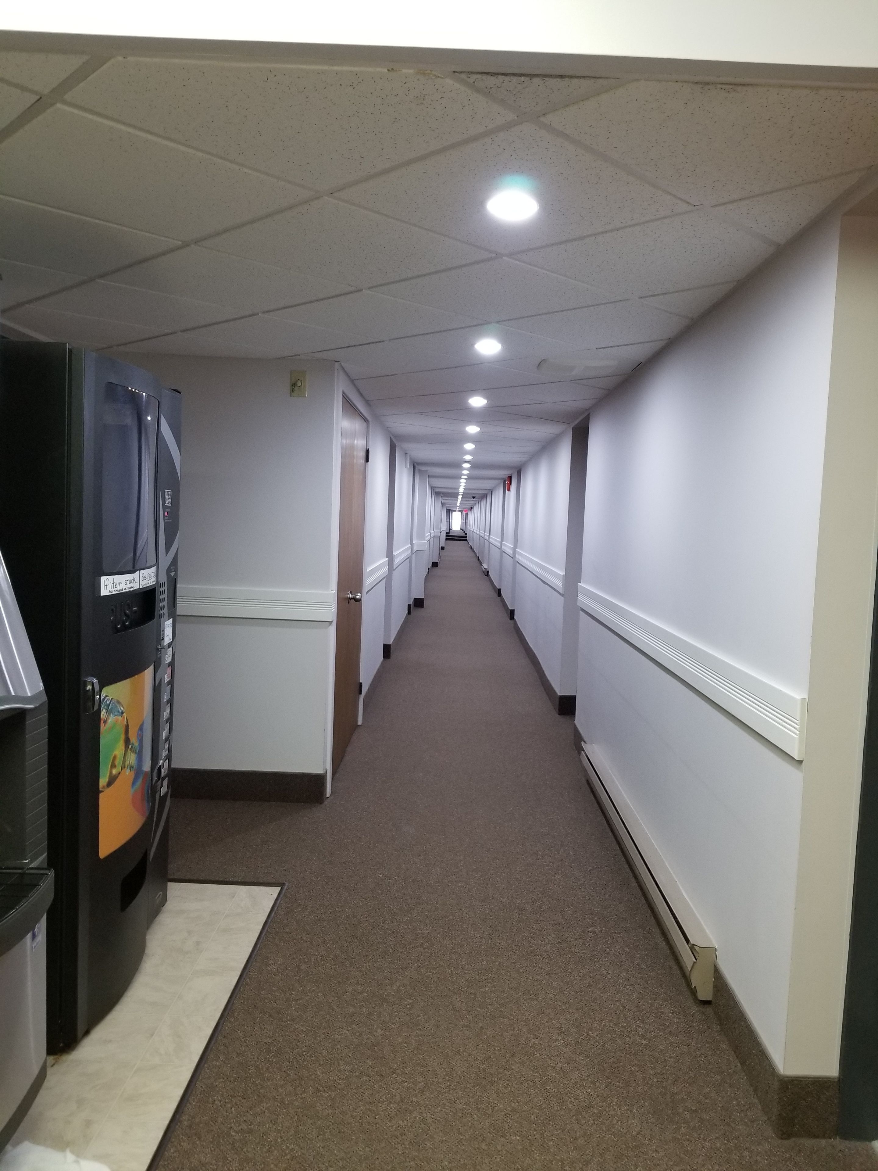 A long, narrow hallway with white walls and a carpeted floor, leading to a door at the end.