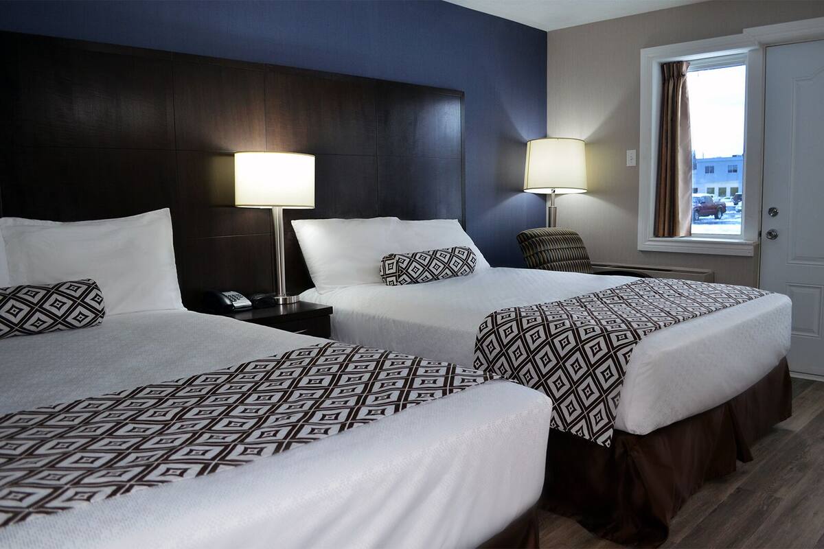 A hotel room with two beds, each with patterned bedding, a dark headboard, bedside lamps, and a window with curtains.