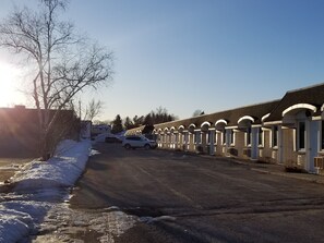 A row of single-story buildings with arched doorways, a paved driveway, and parked cars.