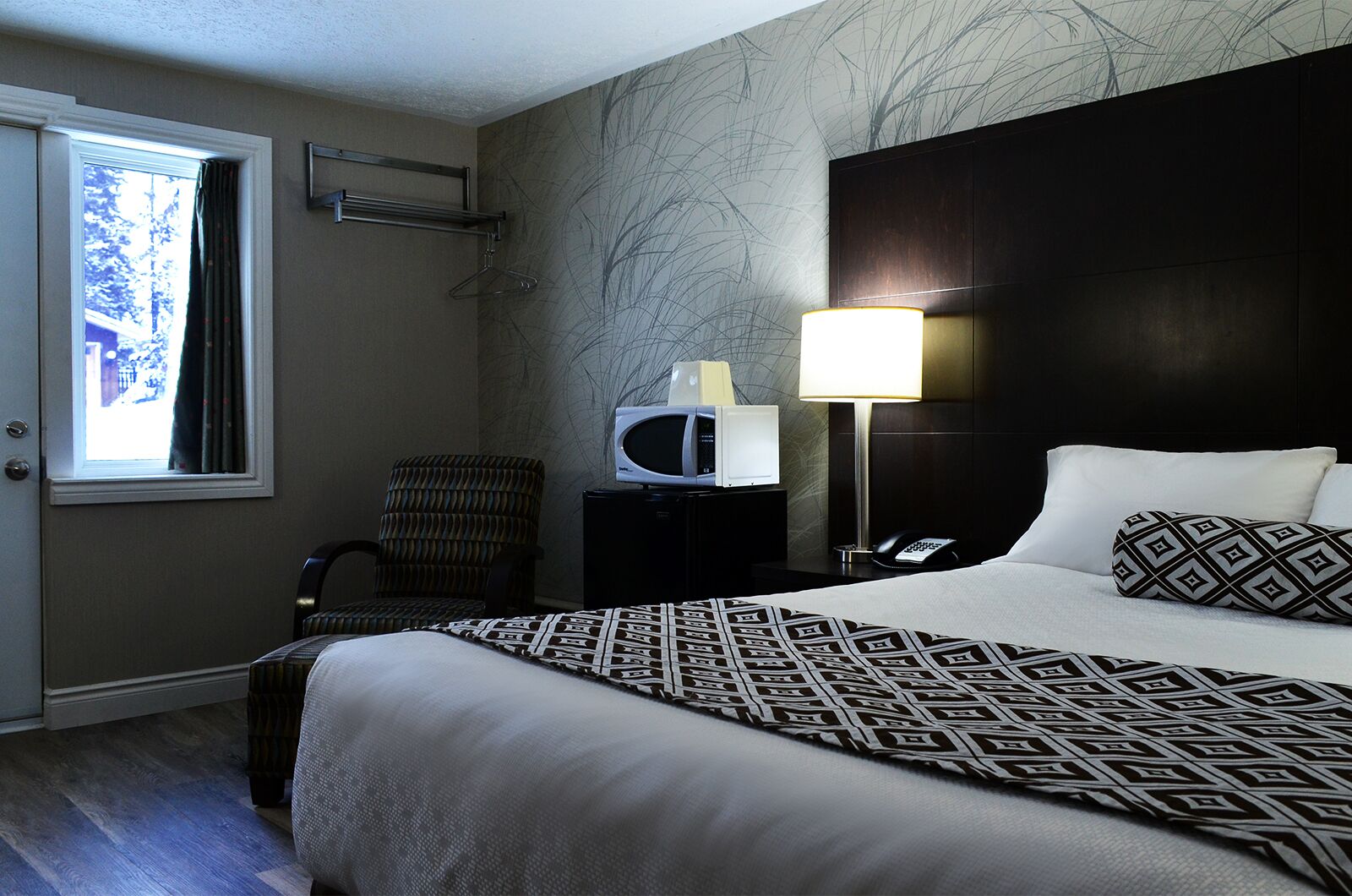A hotel room with a bed, a chair, a desk with a lamp, a telephone, and a window.