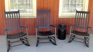 Three wooden rocking chairs with metal frames and slatted seats, positioned in front of a wooden wall with two windows. - The Chepstow Inn (Brockton)
