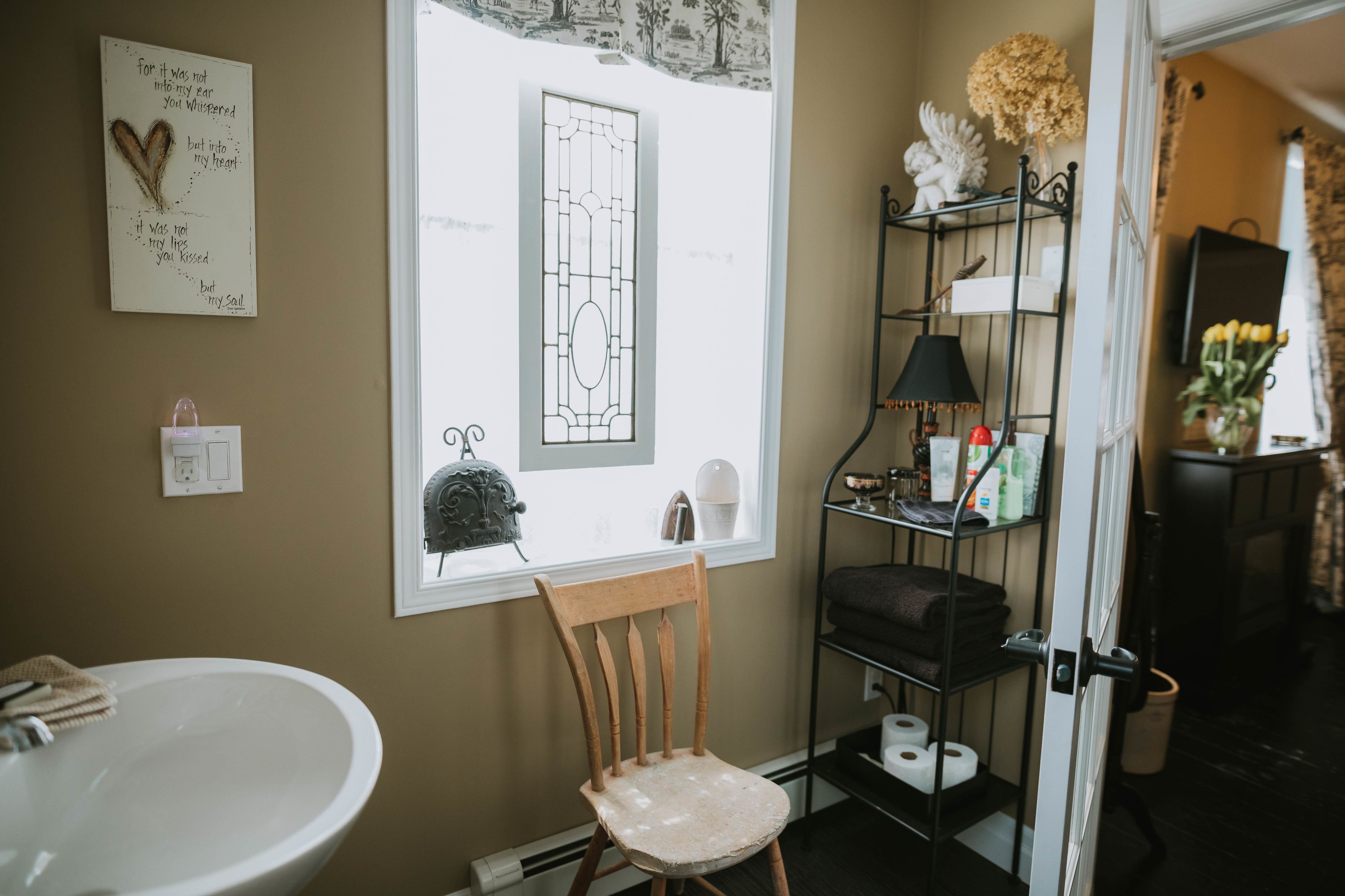 A bathroom with a white sink, a wooden chair, a shelf with toiletries, and a window with a view of a house.