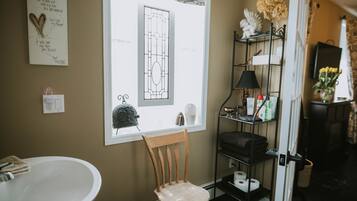 A bathroom with a white sink, a wooden chair, a shelf with toiletries, and a window with a view of a house.