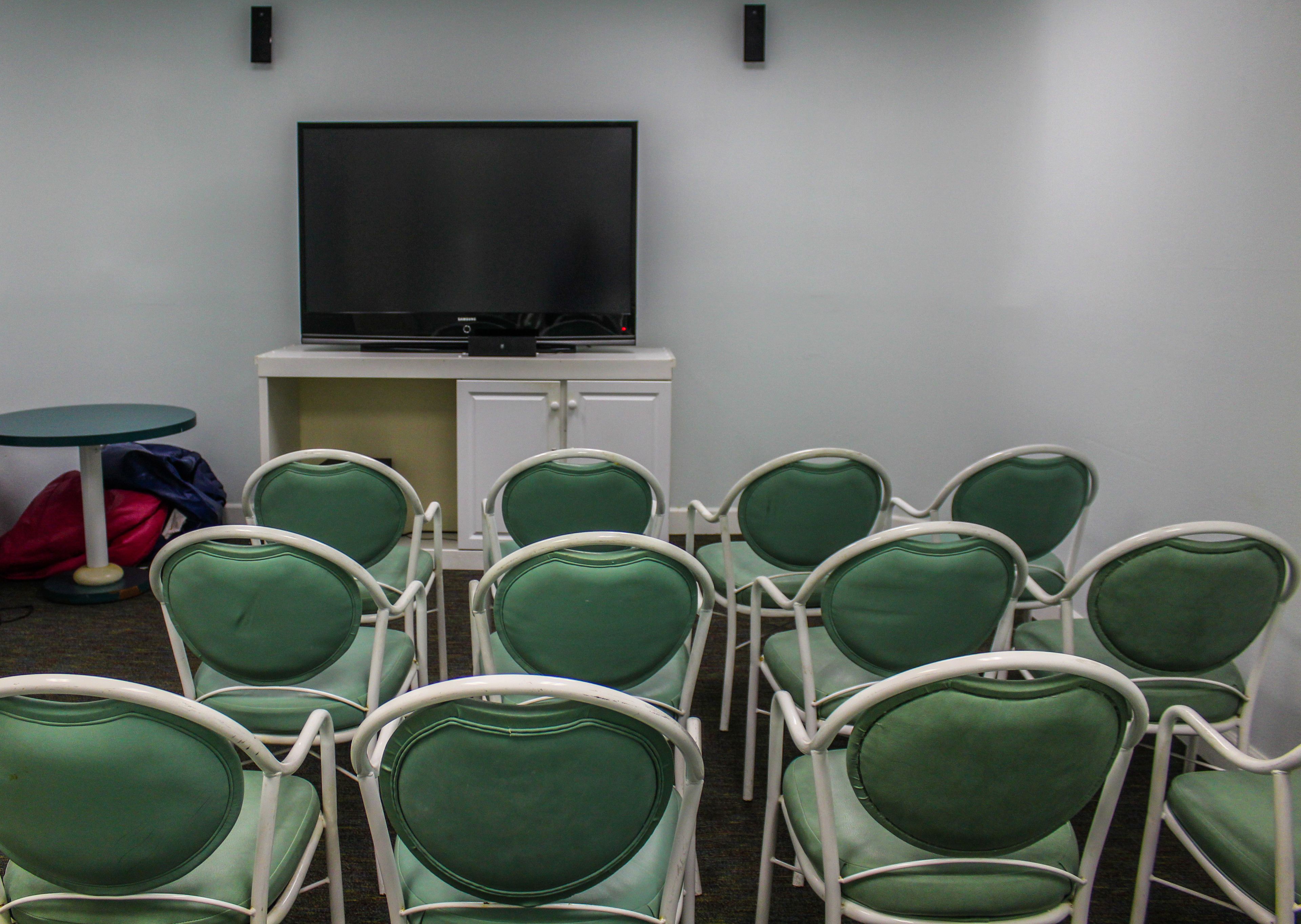 A conference room with green chairs arranged in rows, a flat-screen TV on a stand, and a small table with a red bag on it.