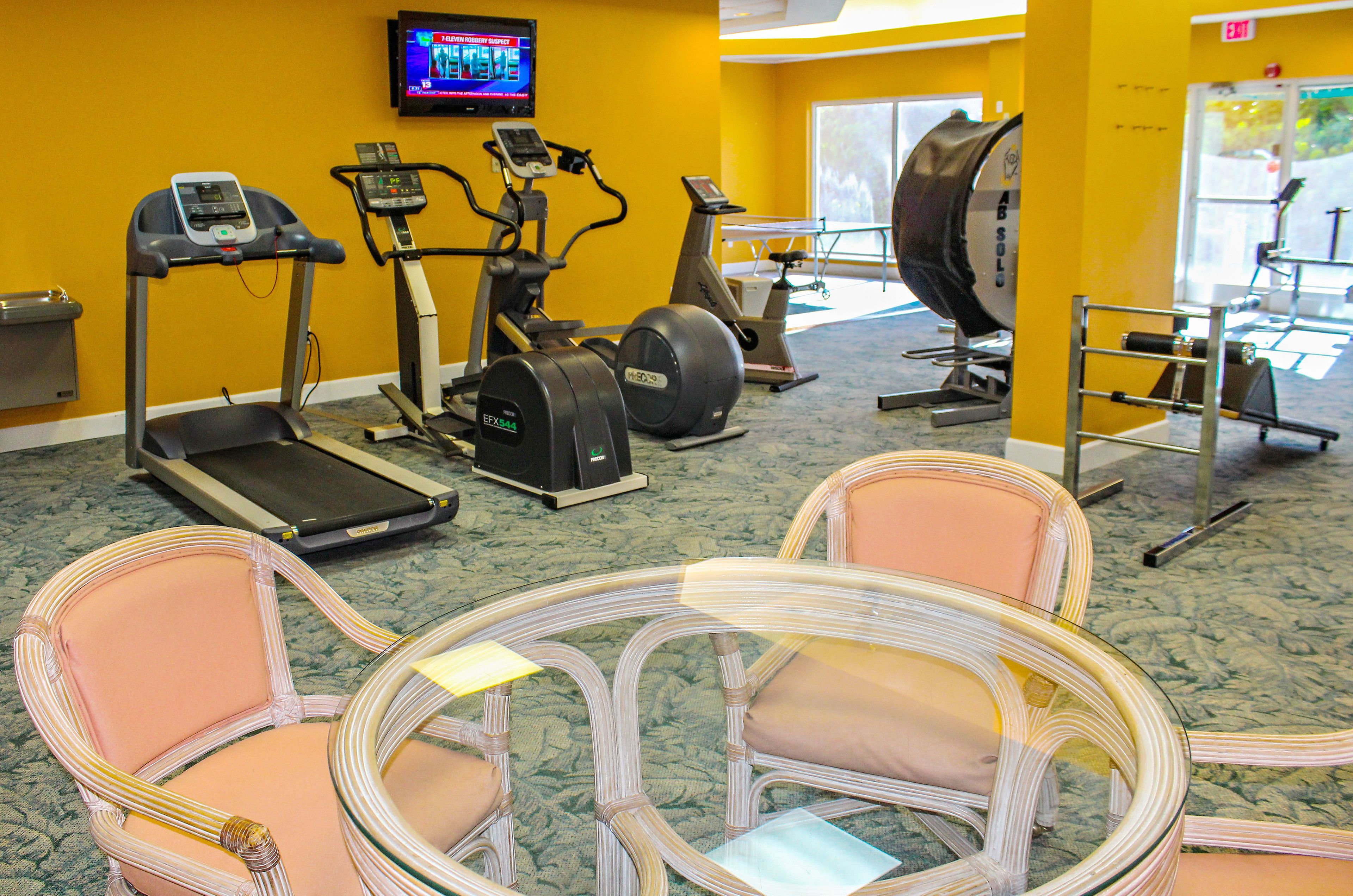 A gym with treadmills, elliptical machines, and a stationary bike. There are wicker chairs and a television mounted on the wall.