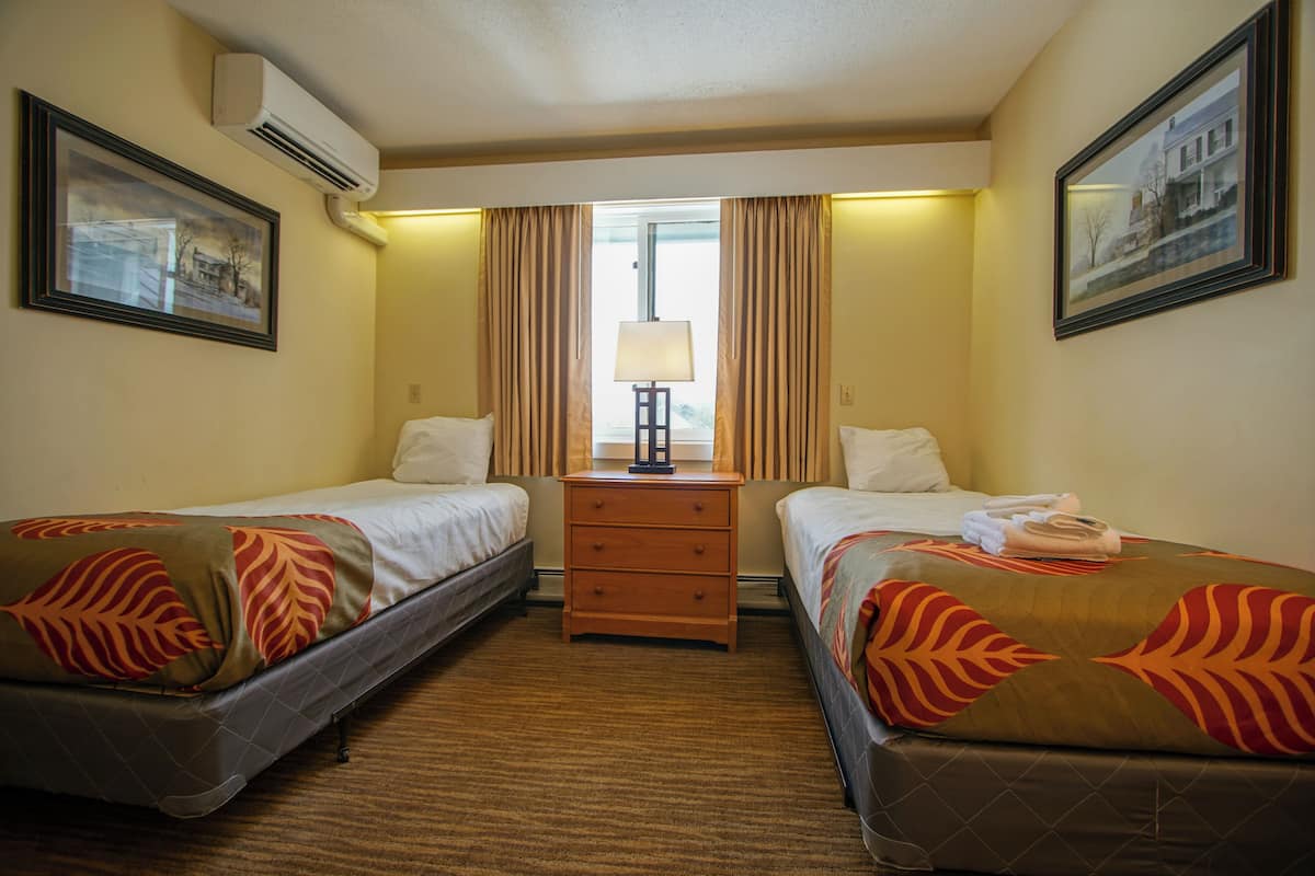 A hotel room with two beds, a wooden dresser, a lamp, and framed pictures on the walls.
