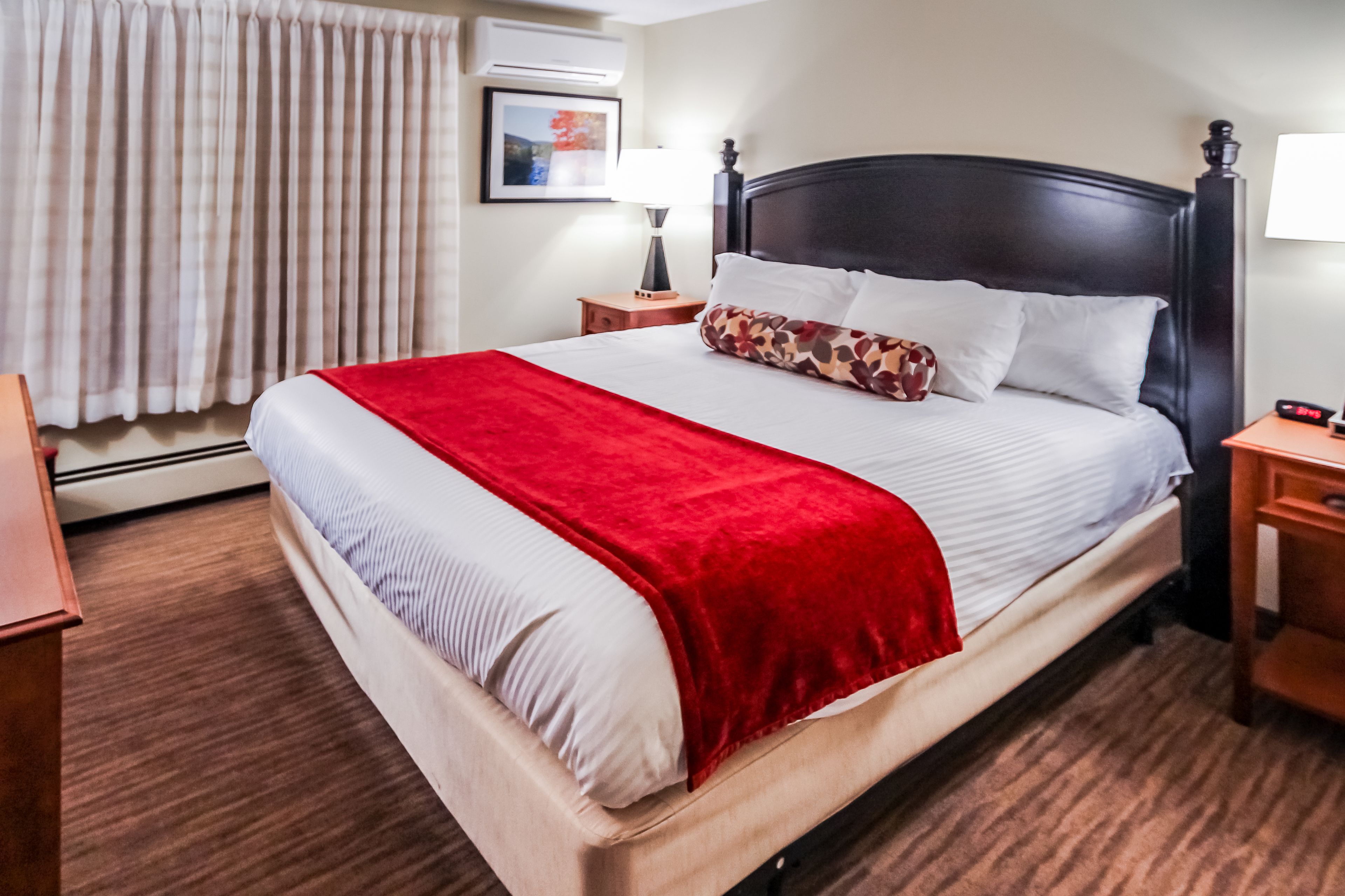 Ein ordentlich bezogenes Bett mit roter Decke, einem Holzkopfschild und einem Nachttisch mit Lampe.