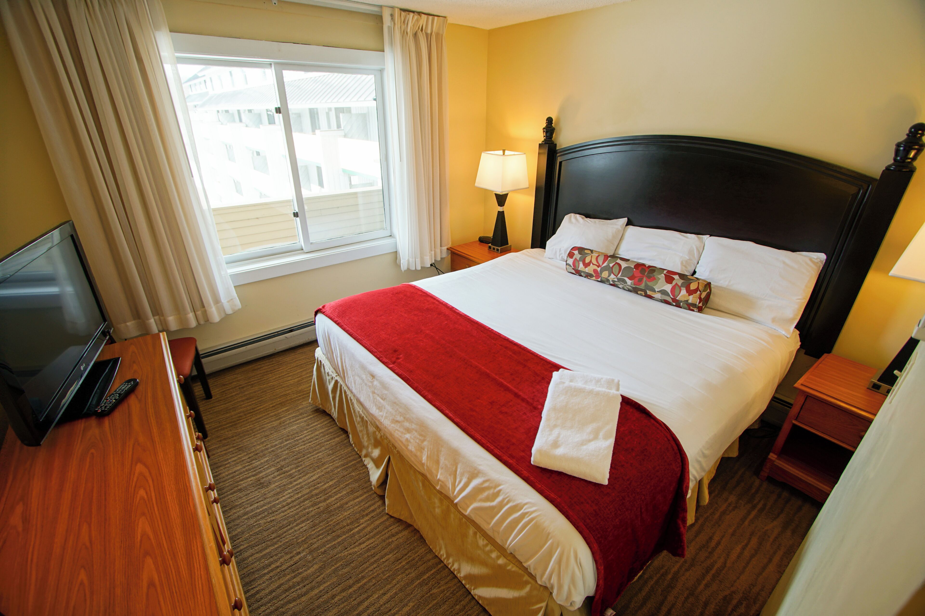 A hotel room with a bed, a desk with a laptop, a window with curtains, and a nightstand with a lamp.