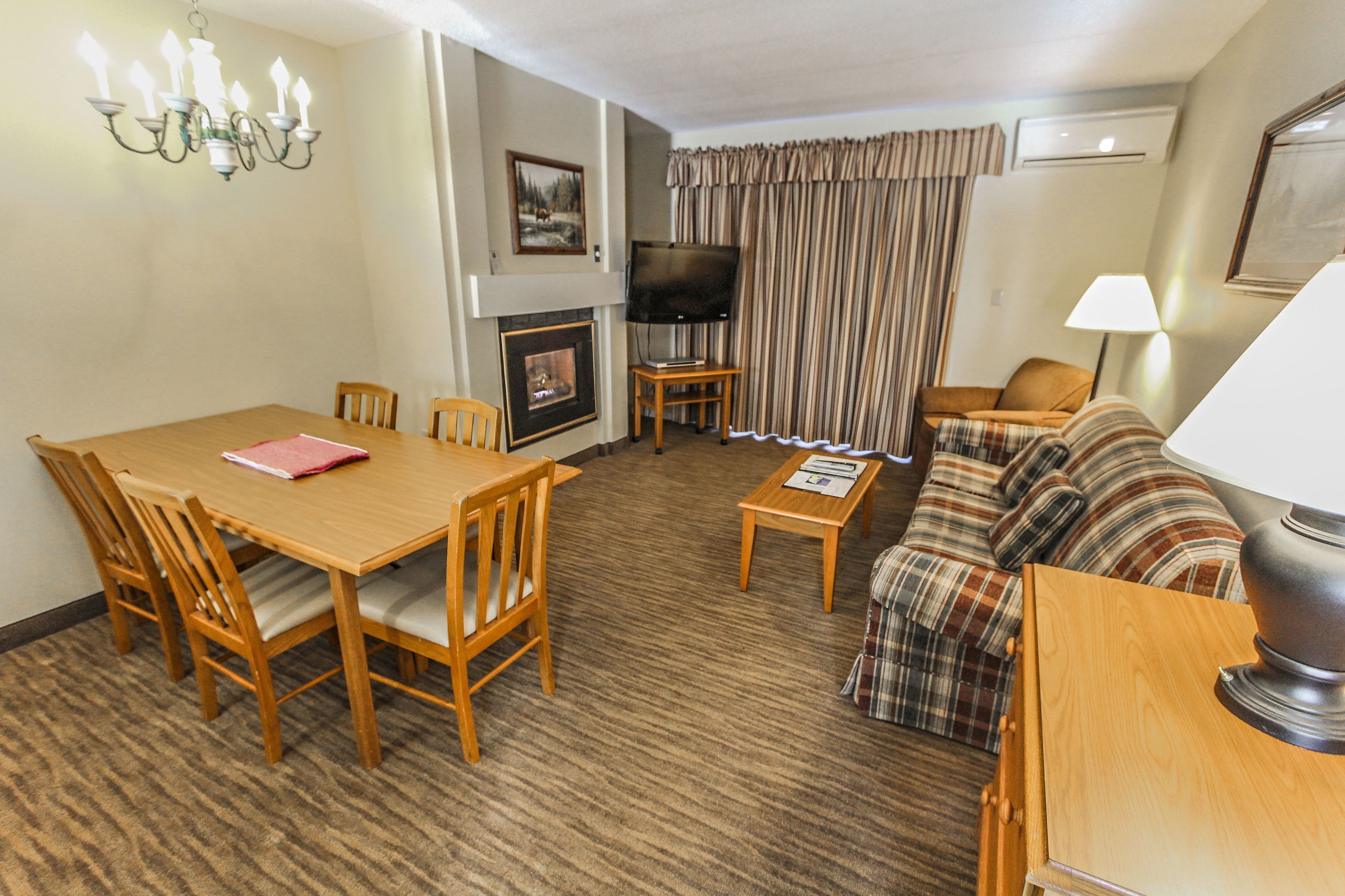A hotel room with a wooden dining table, chairs, a sofa, a TV, a fireplace, and a lamp.