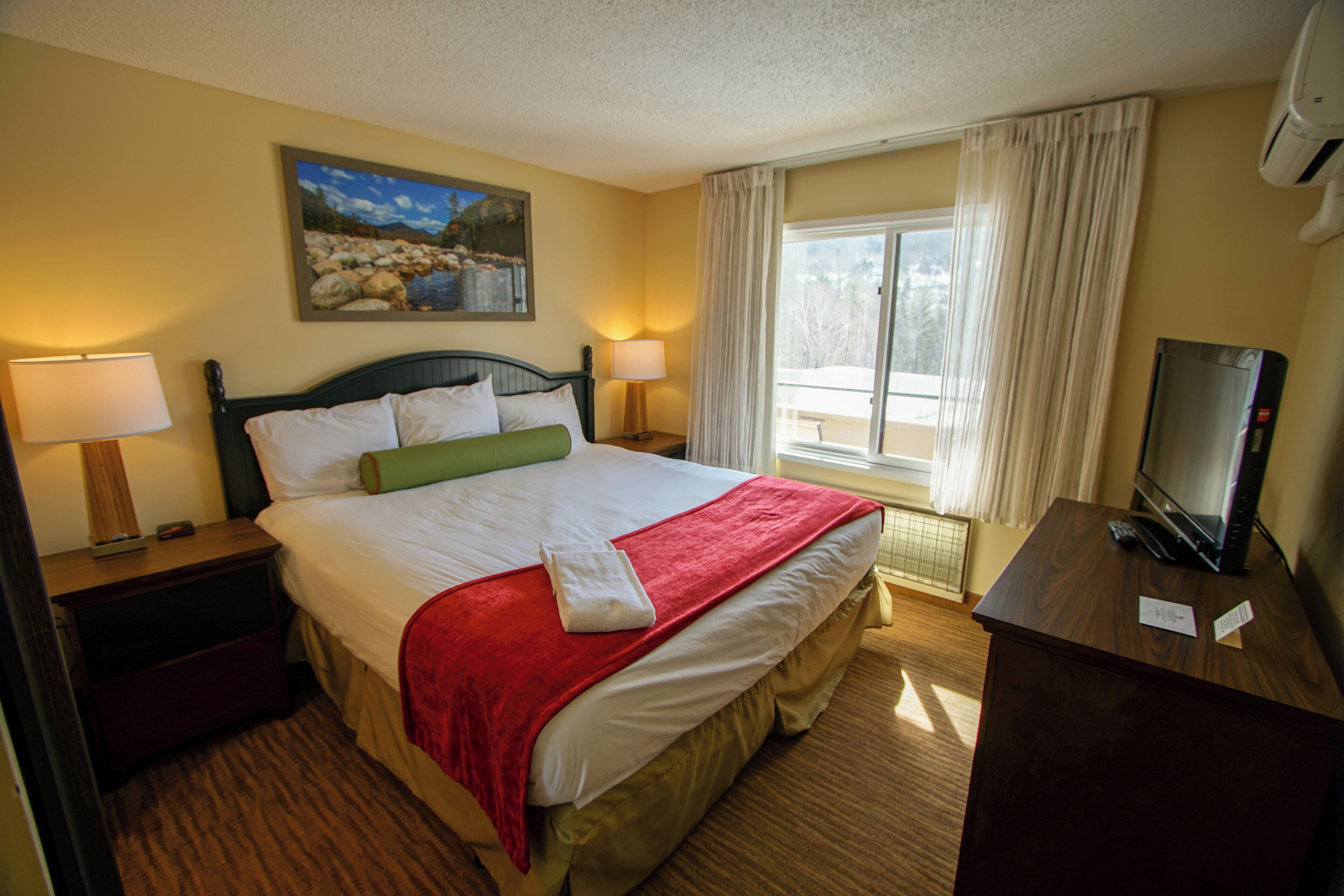 A hotel room with a bed, two bedside tables with lamps, a TV, a painting on the wall, and a window with curtains.