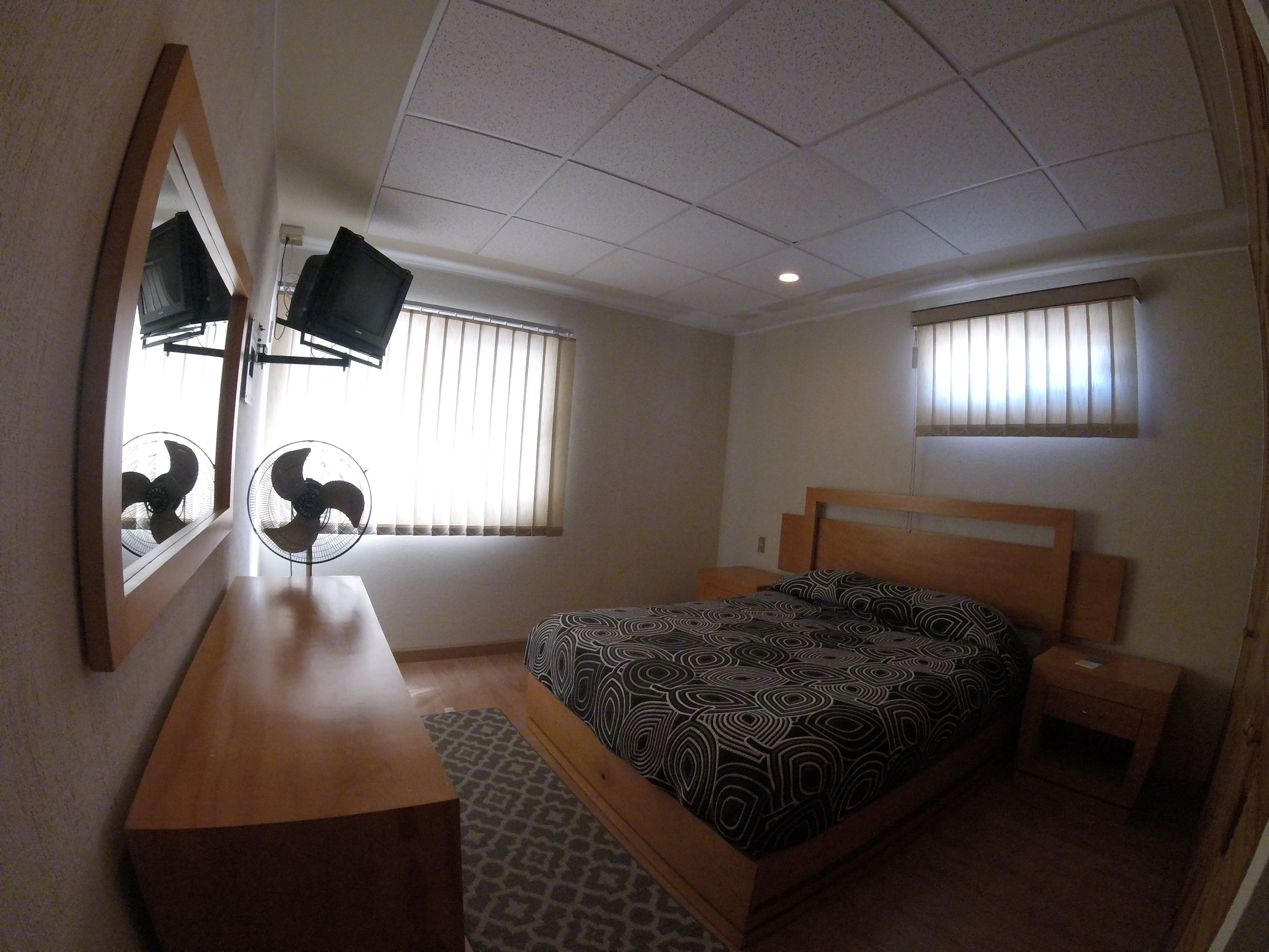 Un dormitorio con cama, mesitas de noche, cómoda, espejo y dos ventanas con persianas.