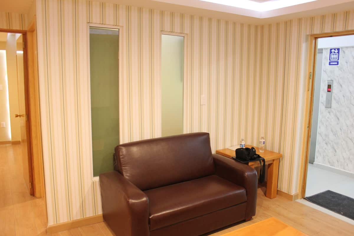 Une pièce avec un canapé en cuir marron, une table d’appoint en bois sur laquelle se trouvent une bouteille et un sac, un papier peint à rayures, une porte avec un miroir et un ascenseur doté d’un bouton.