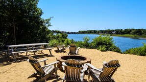 A beachside seating area with Adirondack chairs and a picnic table, overlooking a calm lake. - The Cove at Yarmouth Resort (West Yarmouth)