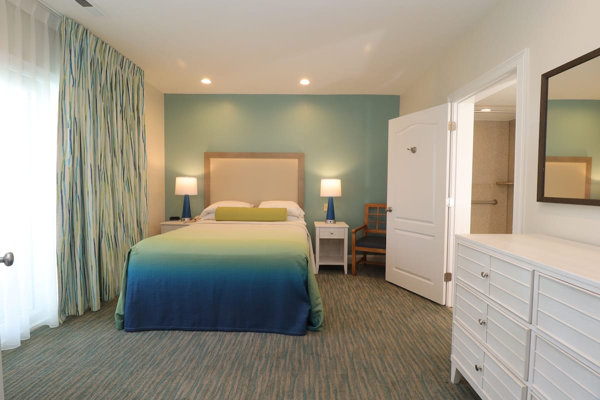 Quarto de hotel com cama, mesas de cabeceira, cômoda, cadeira e porta para o banheiro.
