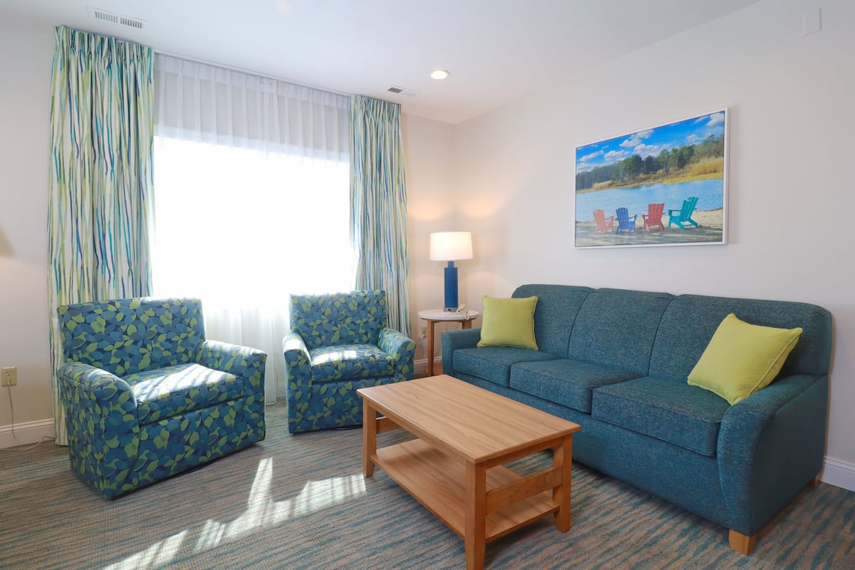 Uma sala de estar com um sofá azul, duas poltronas combinando, uma mesa de centro de madeira e um quadro com cadeiras à beira de um lago.