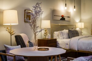 A hotel room with a round dining table, two bedside lamps, a bed with a headboard, a nightstand, and a wall-mounted artwork.