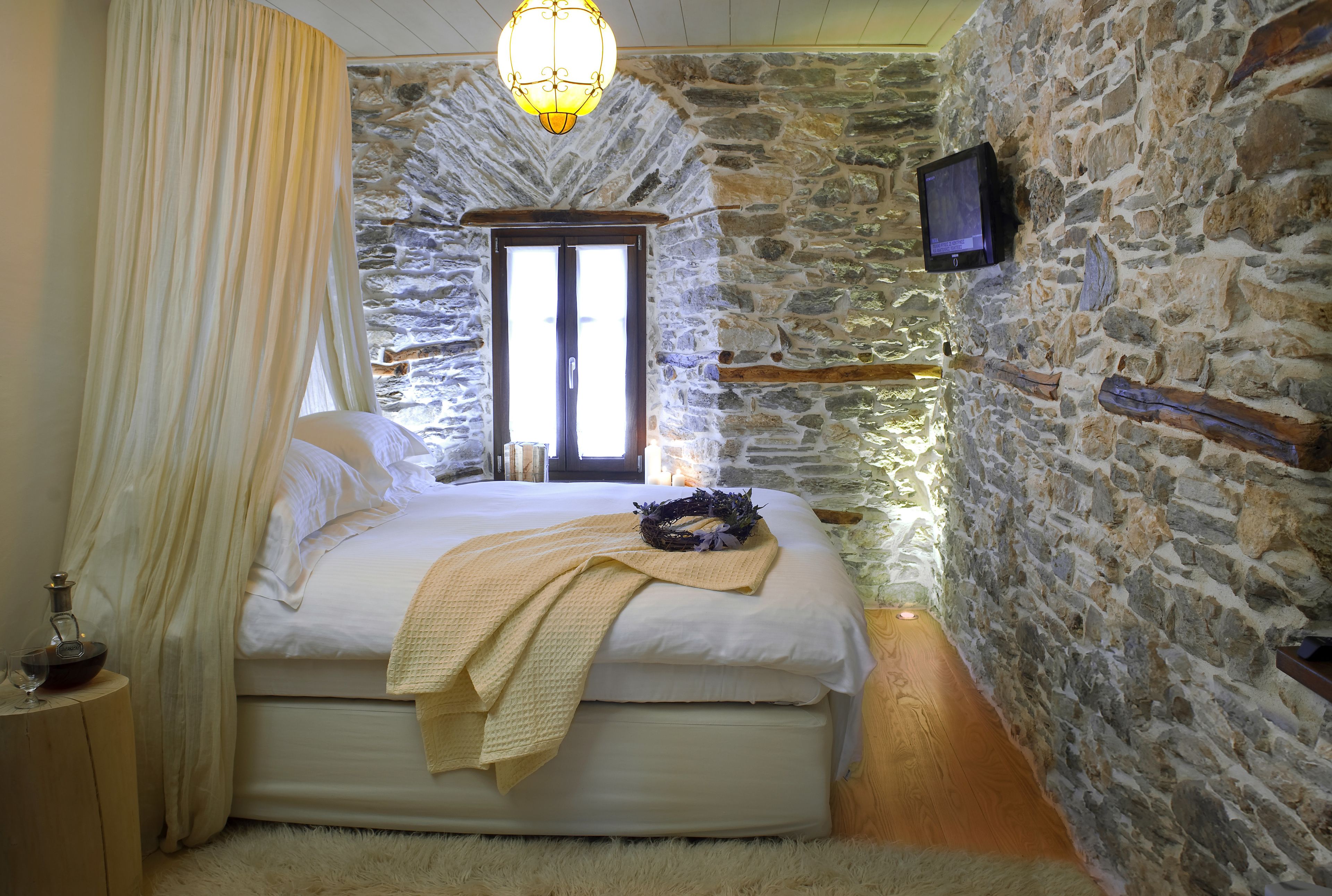A bedroom with a stone wall, a bed with white linens, a wooden headboard, a bedside table with a lamp, and a television mounted on the wall.