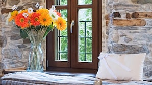 A cozy seating area with a stone wall, a window with a view, a vase of flowers, oranges, and a decanter with wine glasses on a tray. - Mountain's Secret (Zagora-Mouresi)