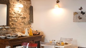 A rustic dining area with stone walls, wooden beams, and a stone fireplace. There are clear glass tables and chairs, and various breakfast items are visible on the tables. - Mountain's Secret (Zagora-Mouresi)