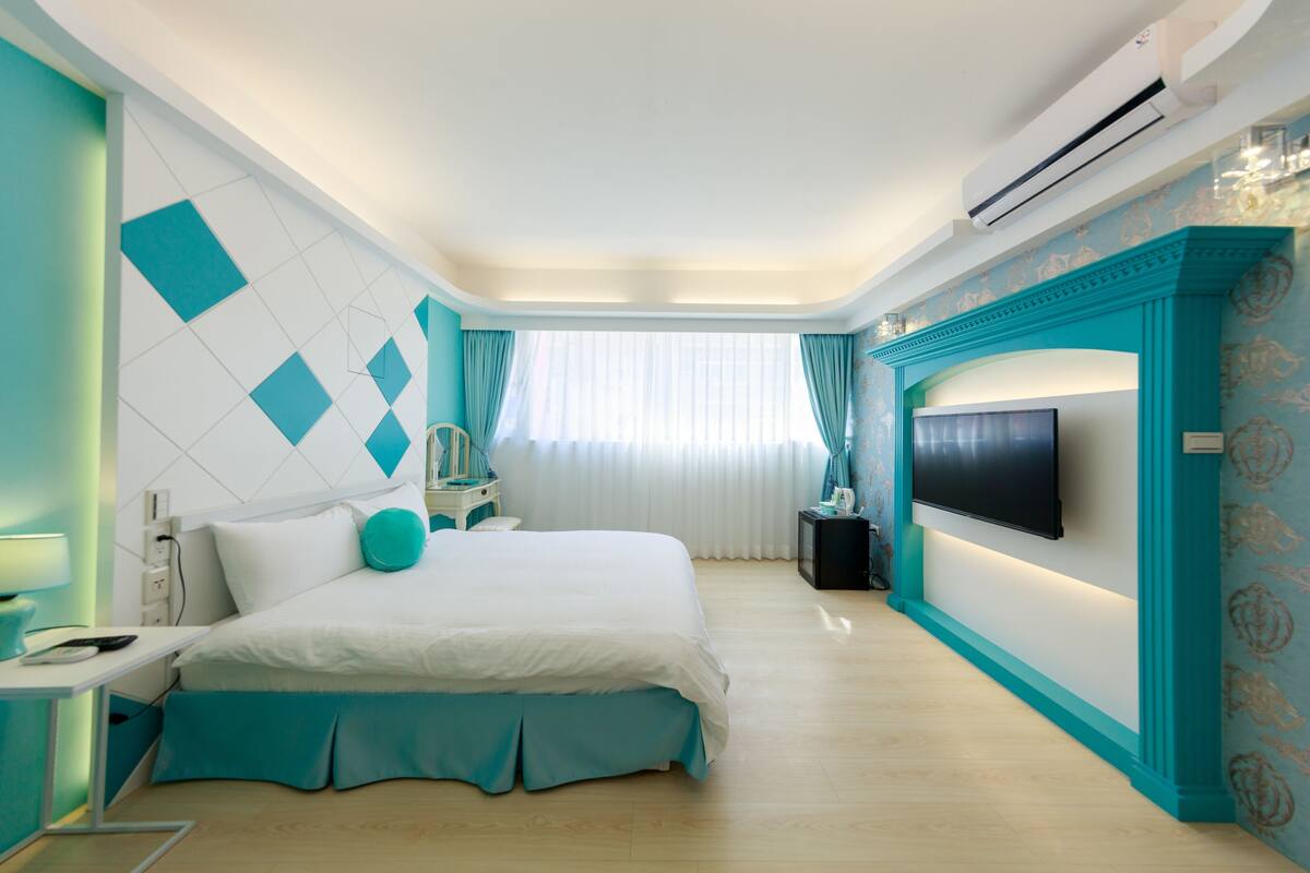 A modern hotel room with a large bed, a flat-screen TV, a bedside table, and a window with curtains.