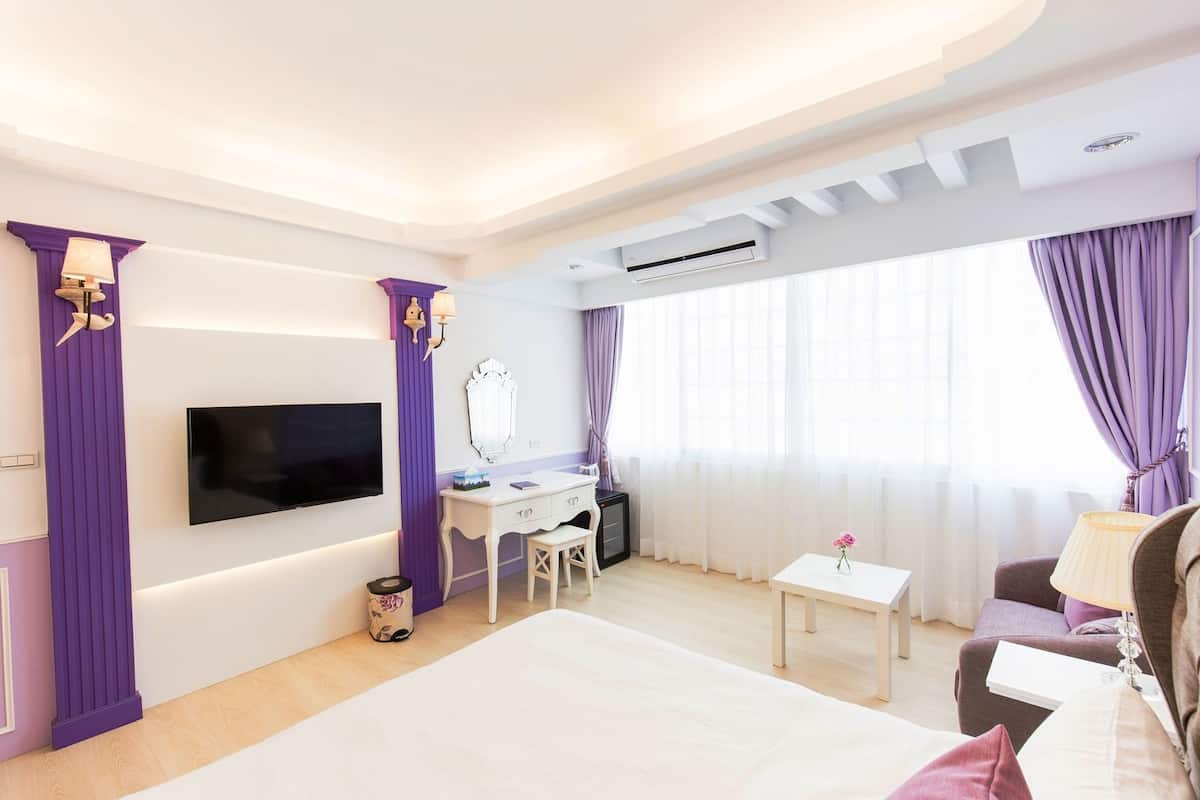 A hotel room with a bed, a desk with a chair, a TV mounted on the wall, an air conditioning unit, and a window with curtains.