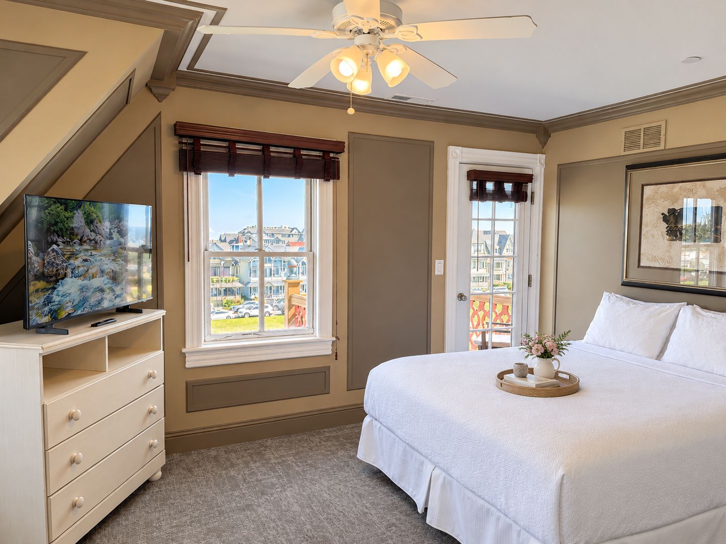Une chambre à coucher comprenant un lit, une commode, un téléviseur, un ventilateur de plafond et une fenêtre donnant sur des maisons.
