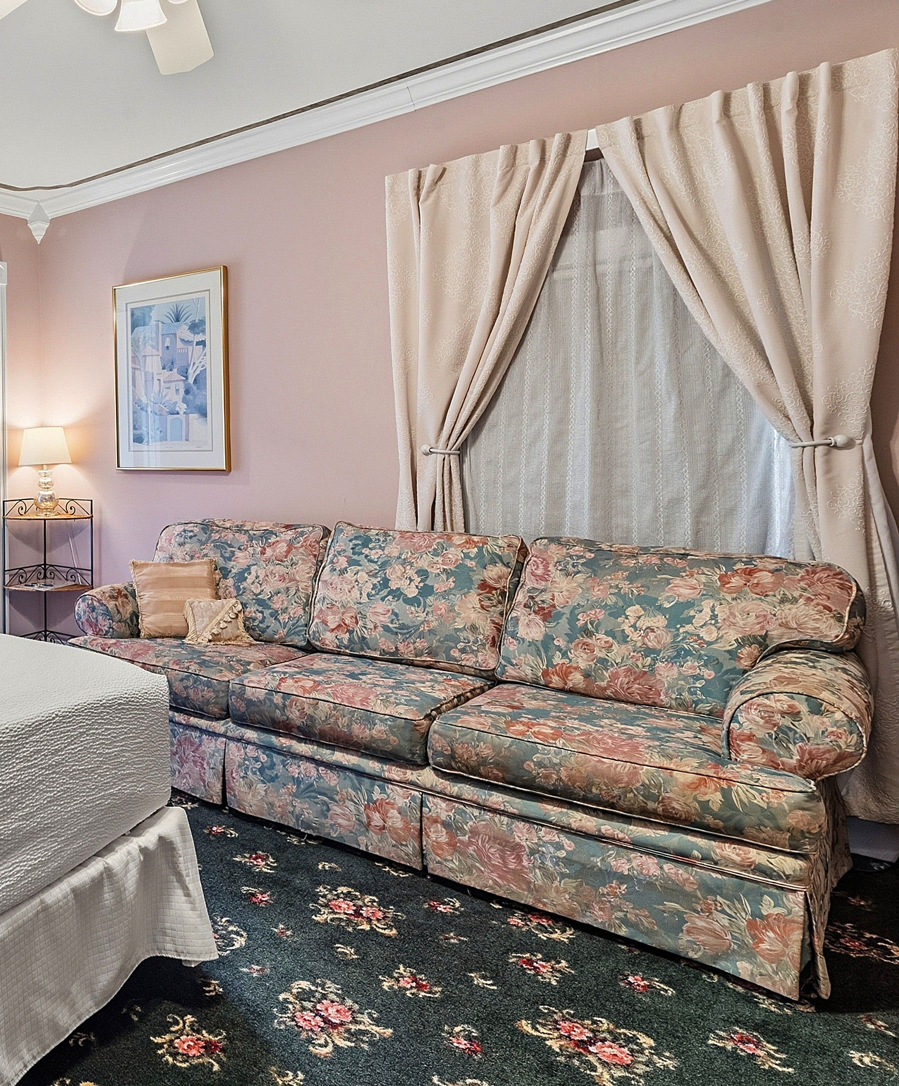 Una habitación con un sofá floral, una cama, una lámpara, un cuadro enmarcado y cortinas.