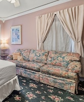 Una habitación con un sofá floral, una cama, una lámpara, un cuadro enmarcado y cortinas.