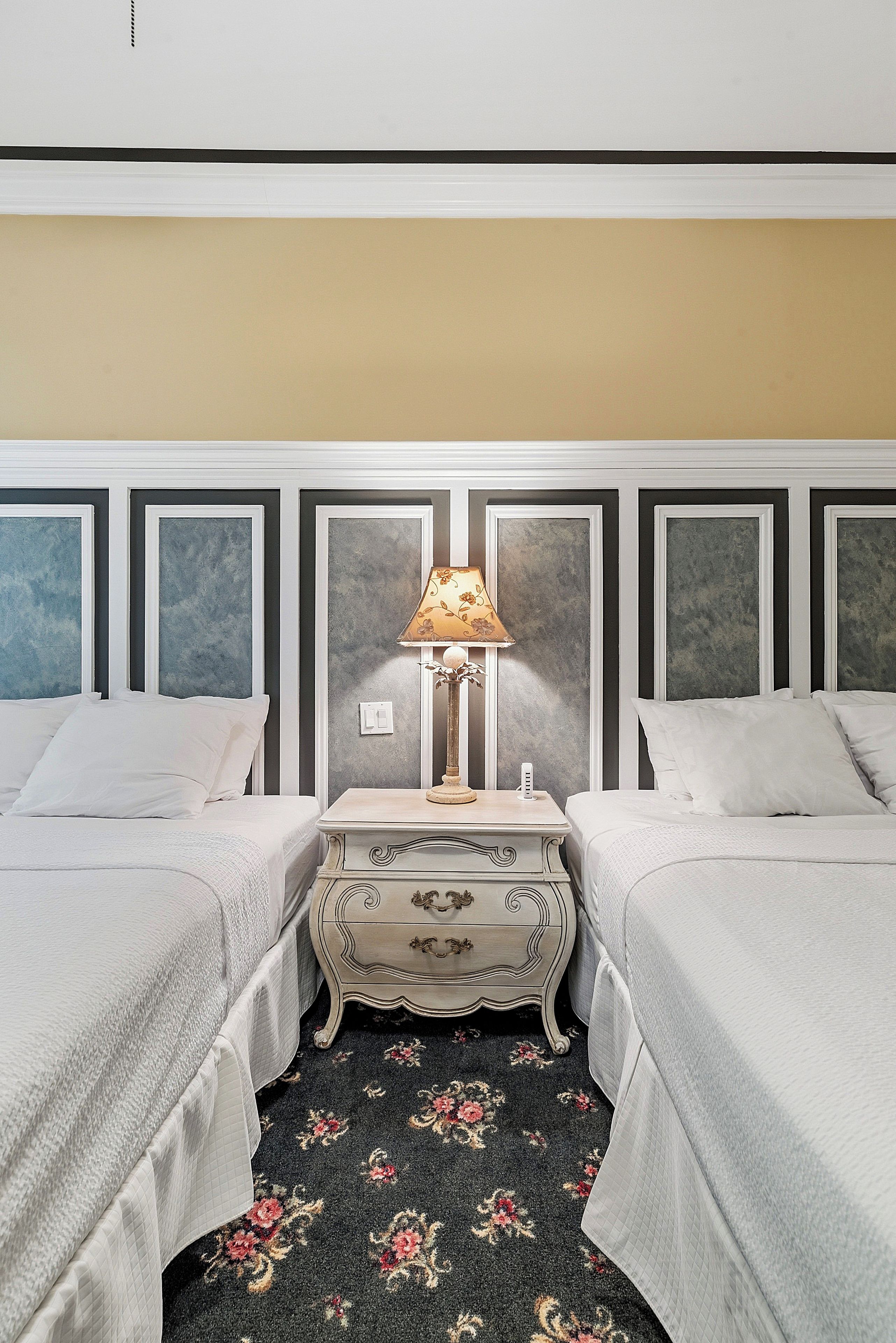 Ein Hotelzimmer mit zwei Betten, einem Nachttisch mit Lampe, einem floralen Teppich und einer schlichten Wand mit Lichtschalter.