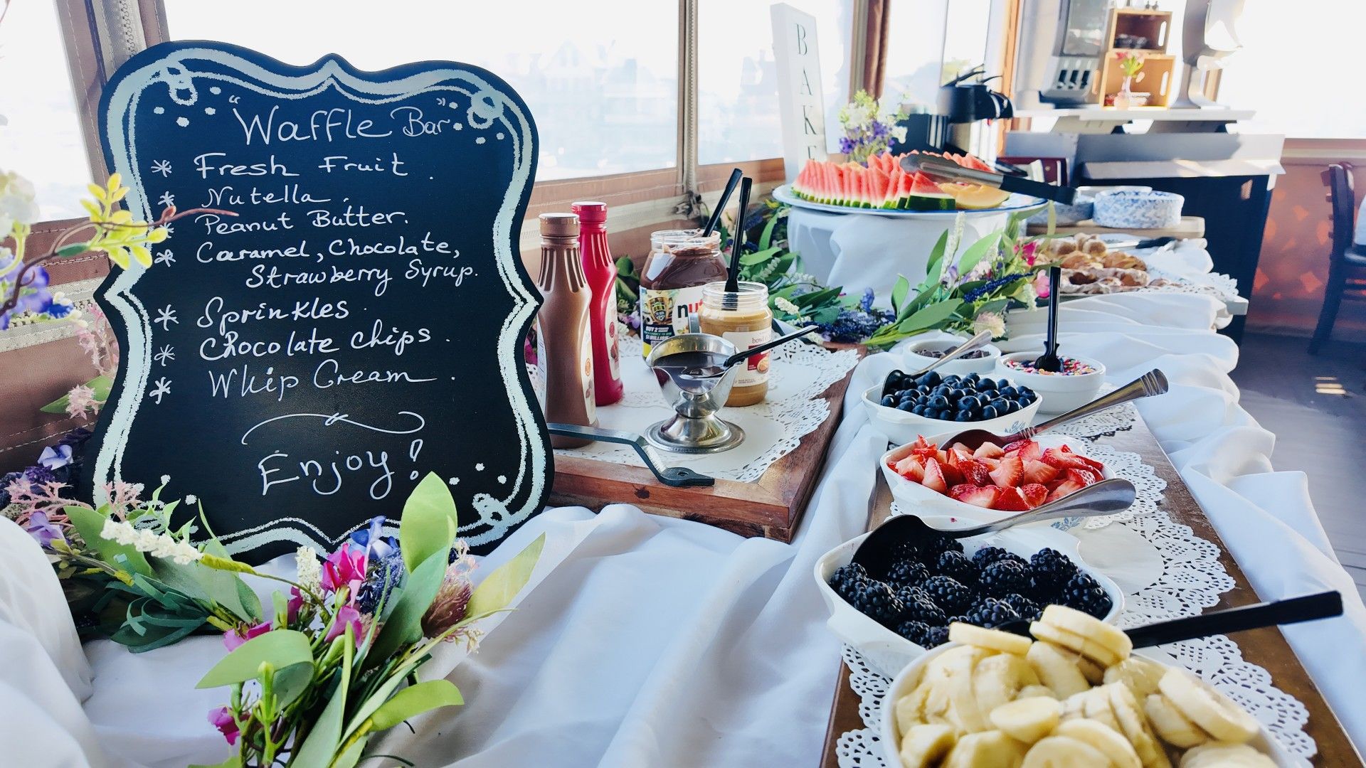 A buffet table with a variety of food items and a chalkboard sign listing offerings such as fresh fruit, nutsella, peanut butter, and chocolate.