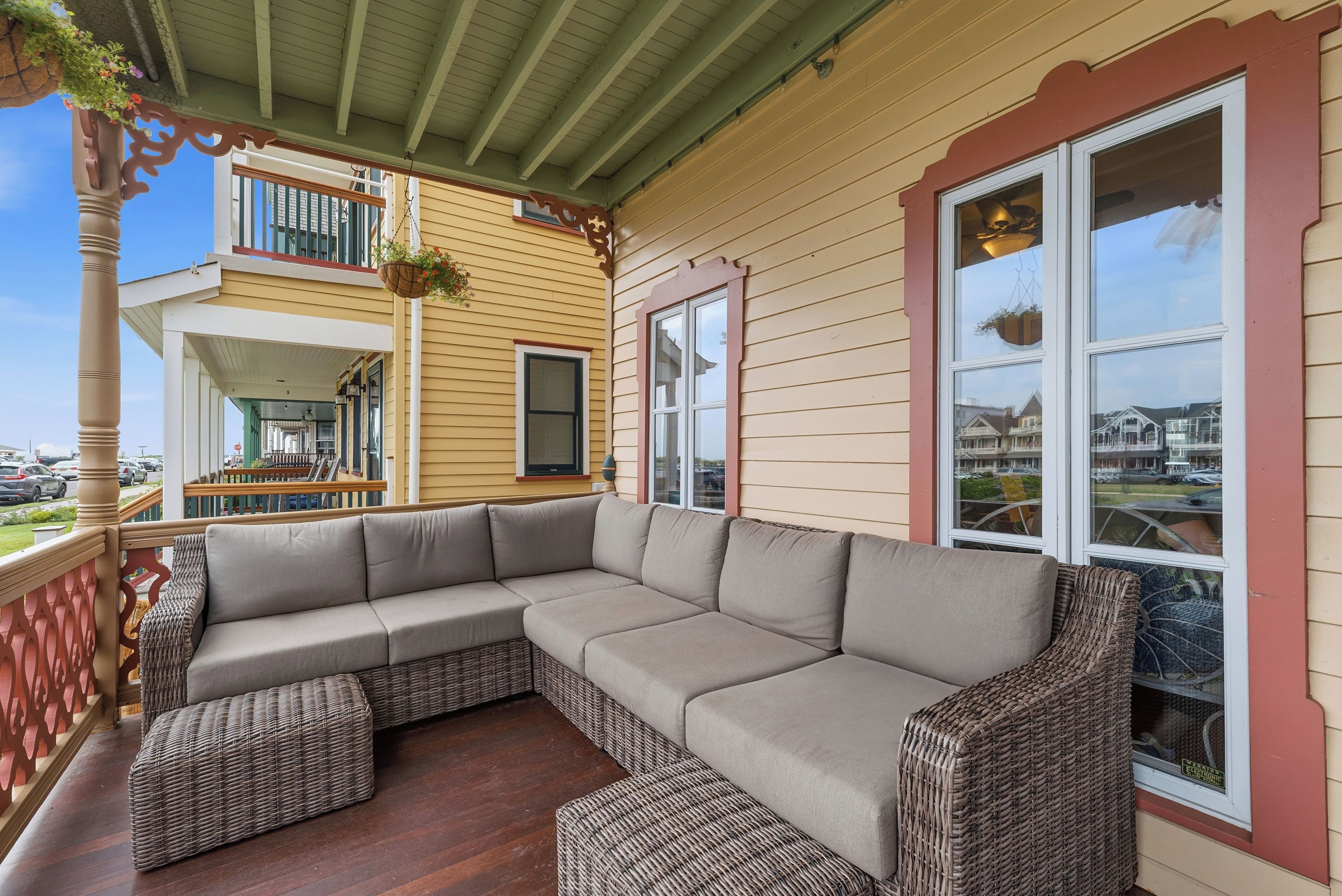 A covered porch with a sectional sofa and wicker ottomans, a balcony with a hanging flower pot, and a view of a residential area.