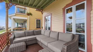 A covered porch with a sectional sofa and wicker ottomans, a balcony with a hanging flower pot, and a view of a residential area.