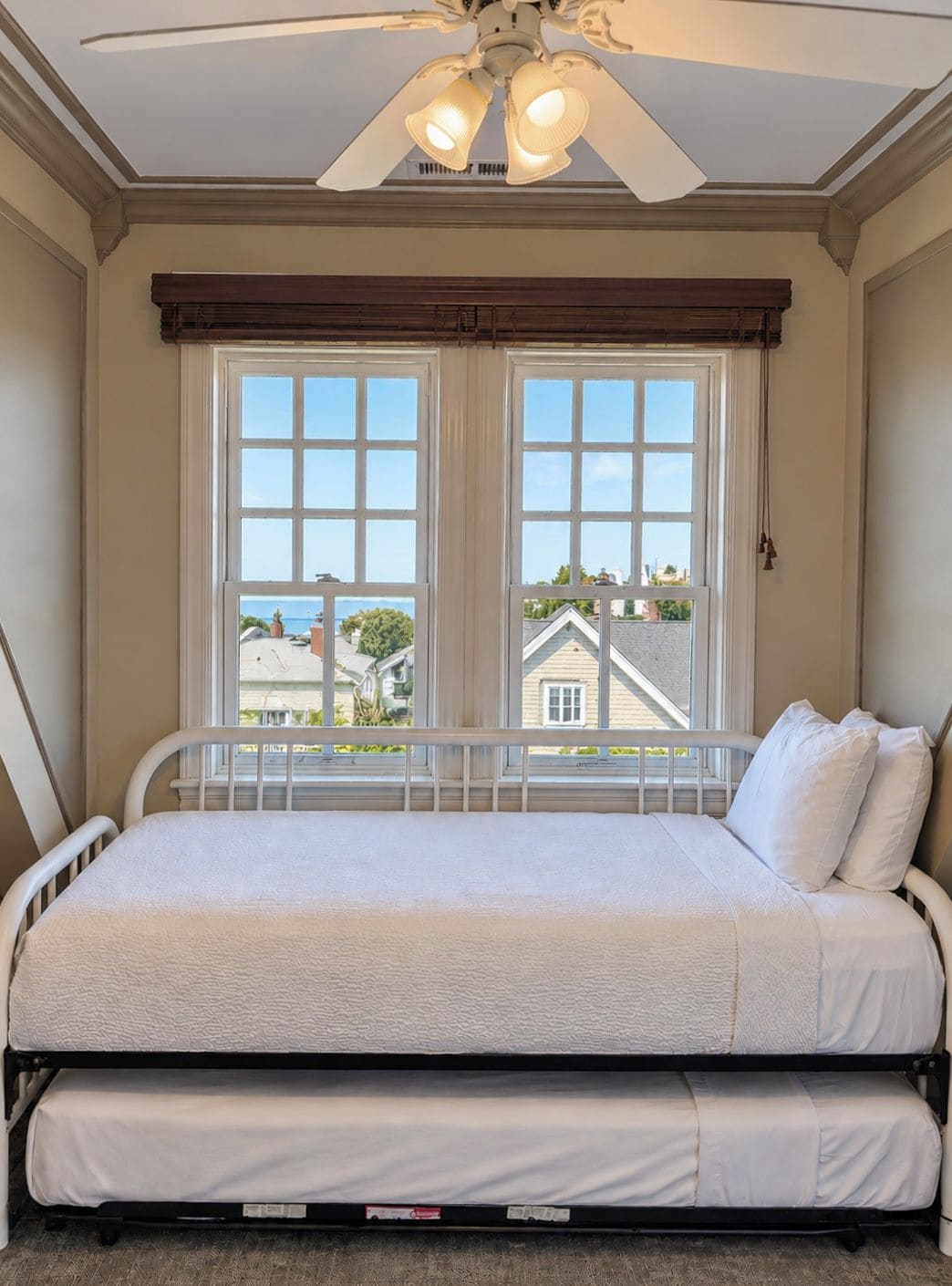 A room with a bed, a ceiling fan, and a window with a view of houses.