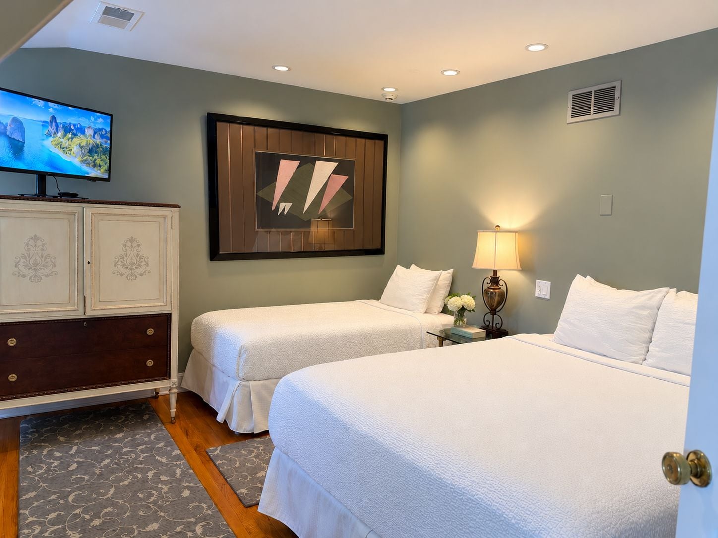 Habitación de hotel con dos camas, televisor, un adorno de pared y una mesita de noche con lámpara.