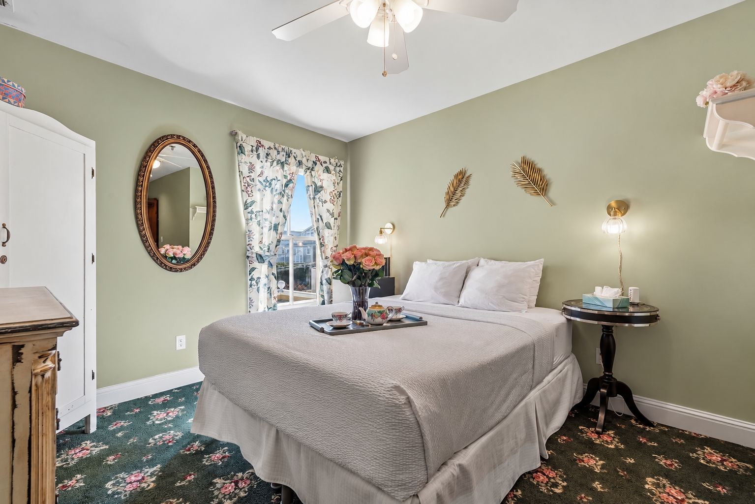 A bedroom with a bed, a bedside table, a mirror, a window with curtains, and a ceiling fan.