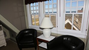 A room with two black leather chairs, a white table with a lamp, and a window with a view of buildings and the ocean. - The Ocean Plaza (Ocean Grove)