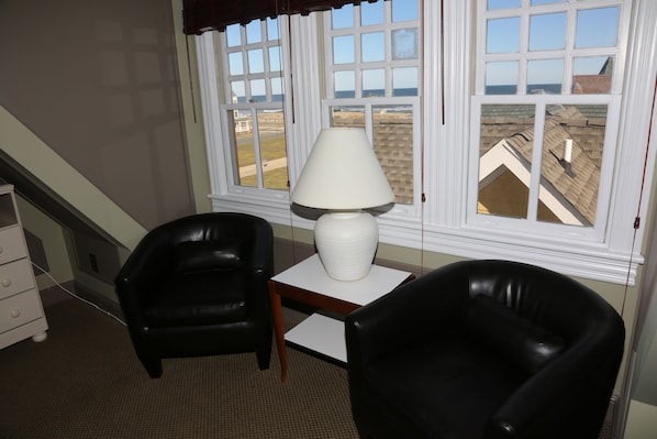 A room with two black leather chairs, a white table with a lamp, and a window with a view of buildings and the ocean. - The Ocean Plaza (Ocean Grove)