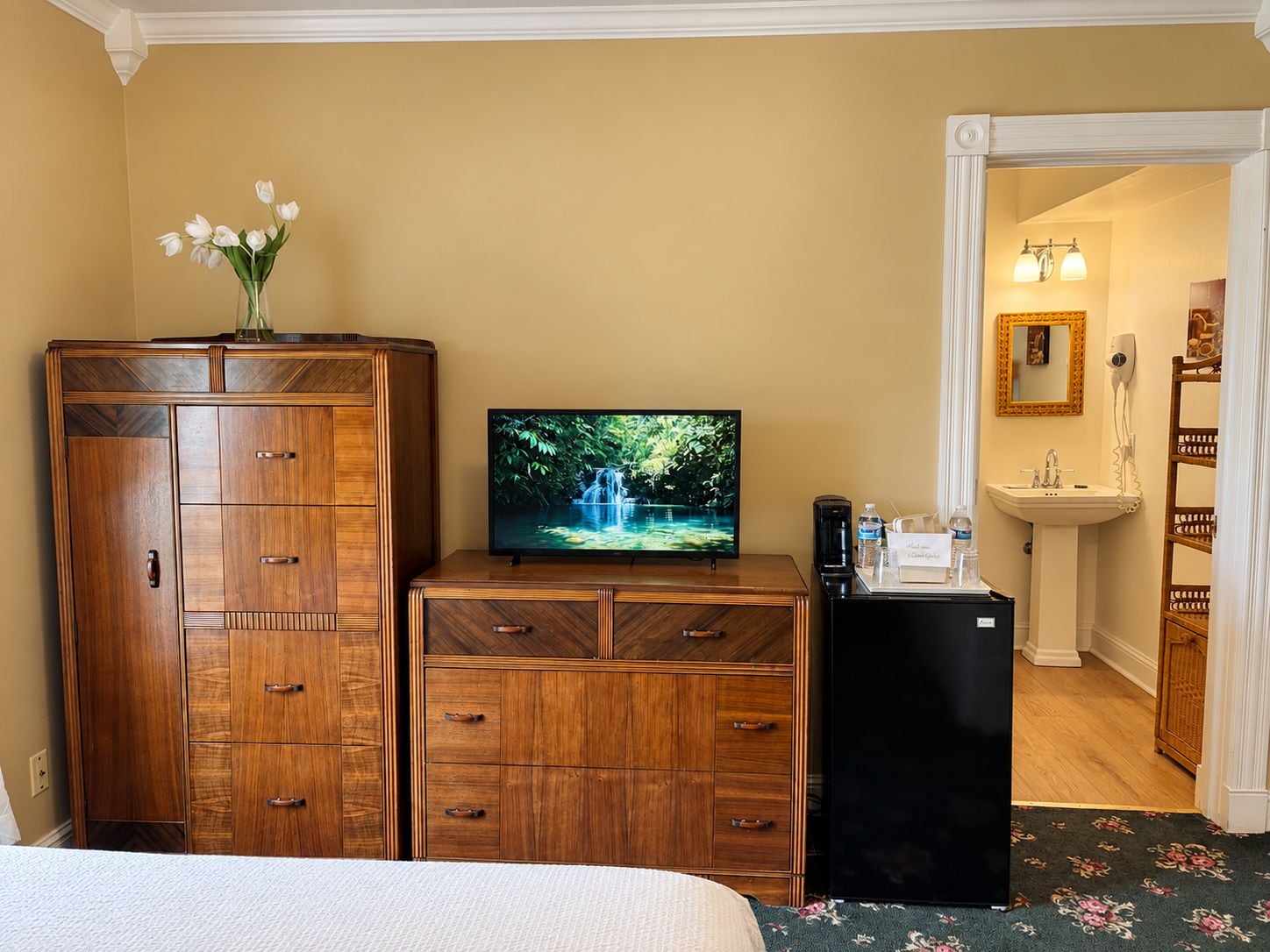 Une chambre d’hôtel avec un lit, une commode en bois, un téléviseur diffusant une image de cascade, un petit réfrigérateur et une salle de bain visible à travers une porte ouverte.