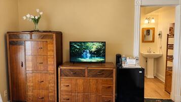 Une chambre d’hôtel avec un lit, une commode en bois, un téléviseur diffusant une image de cascade, un petit réfrigérateur et une salle de bain visible à travers une porte ouverte.