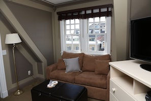 A living room with a brown sofa, a black ottoman, a lamp, and a TV on a white stand. - The Ocean Plaza (Ocean Grove)