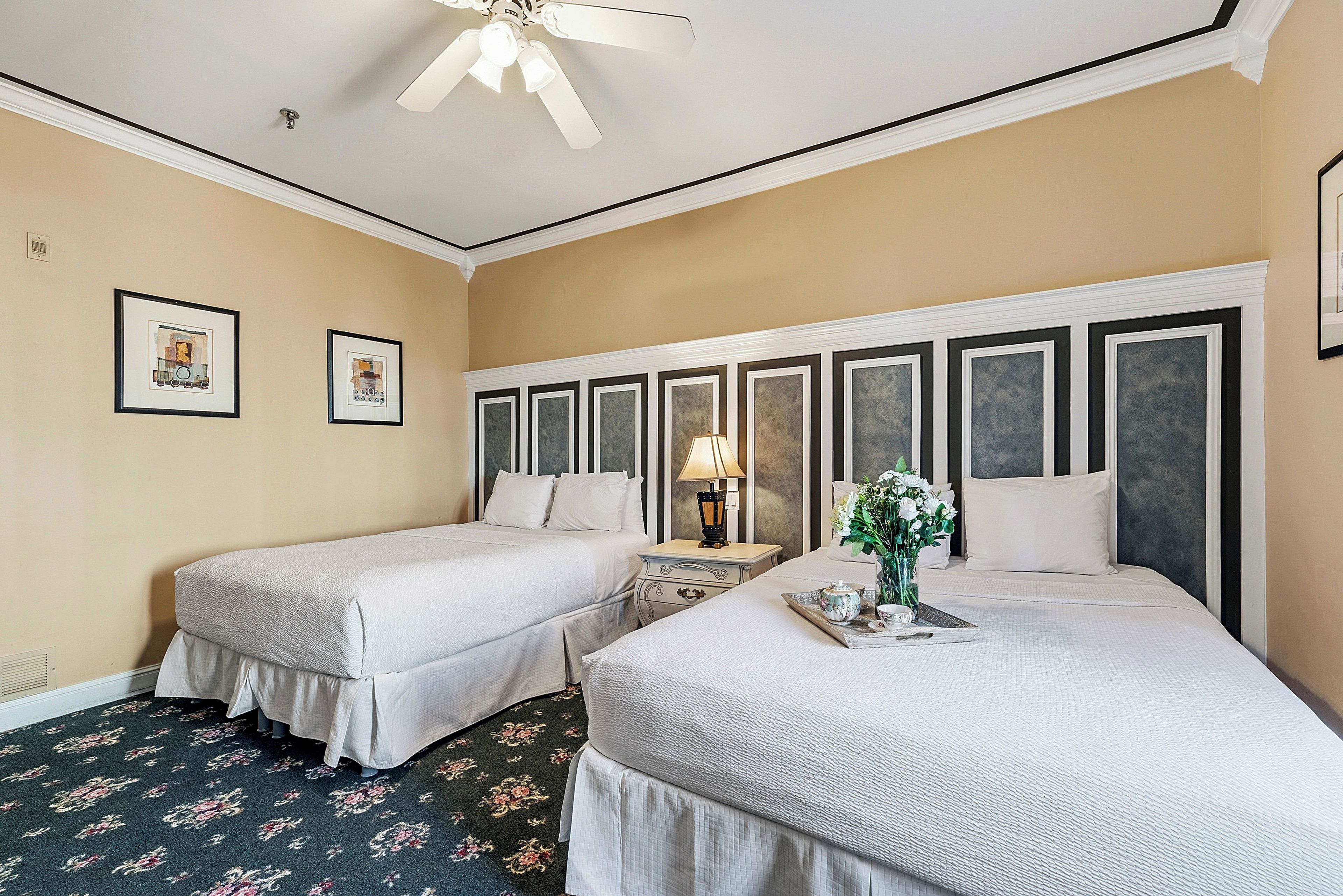Una habitación de hotel con dos camas, un ventilador de techo, obras de arte enmarcadas en la pared y una mesita de noche con una lámpara y una bandeja con un hervidor.