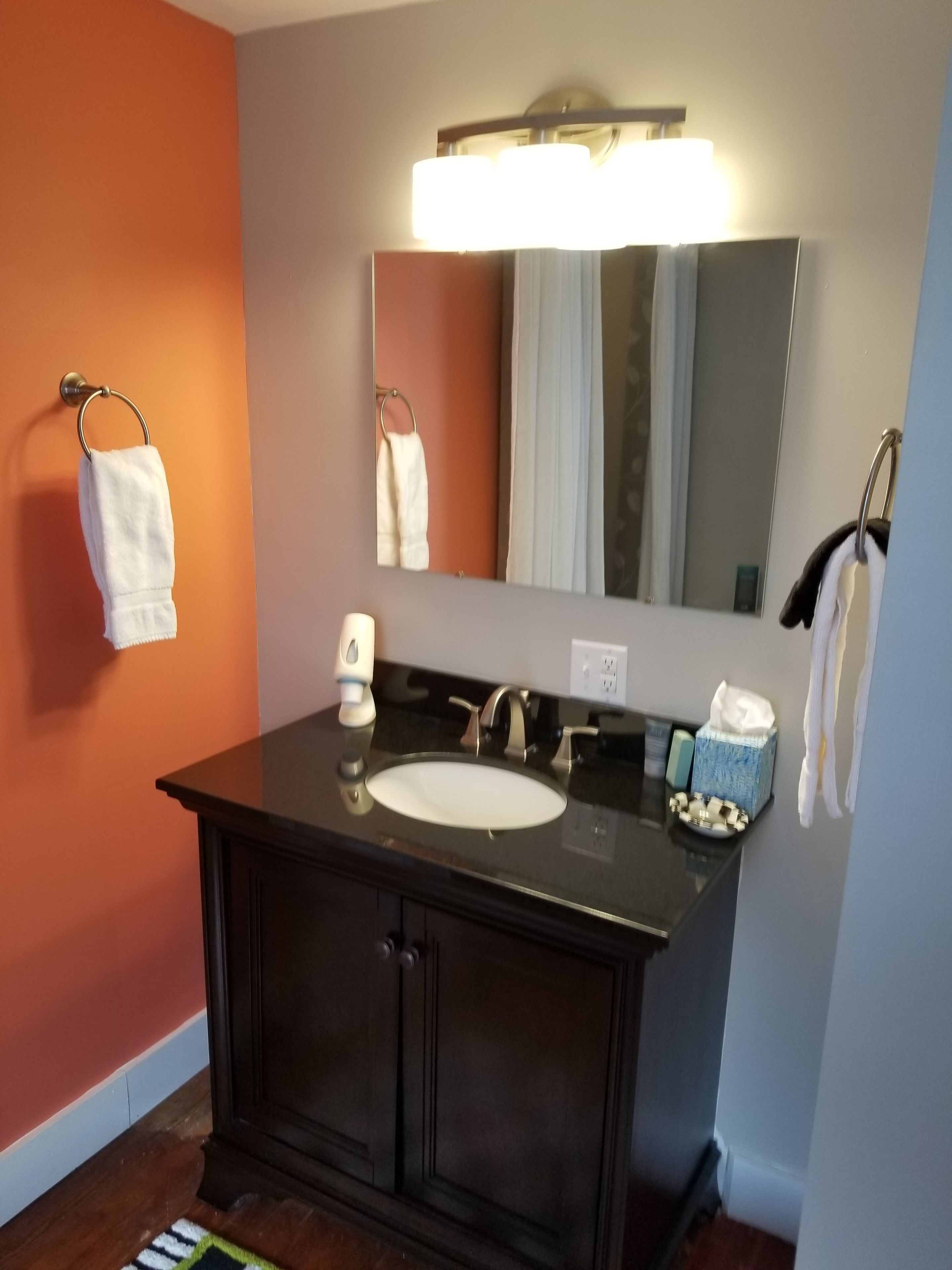 Un baño con tocador oscuro, un lavabo blanco, un espejo y dos toallas colgadas en aros.